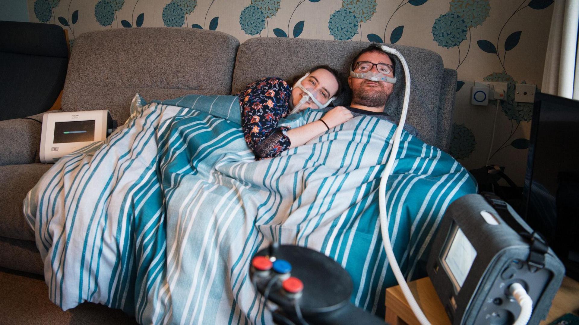 Charlotte and Tom Hardwick sitting on a sofa, covered with a duvet, linked to machines which have a tube running to their noses