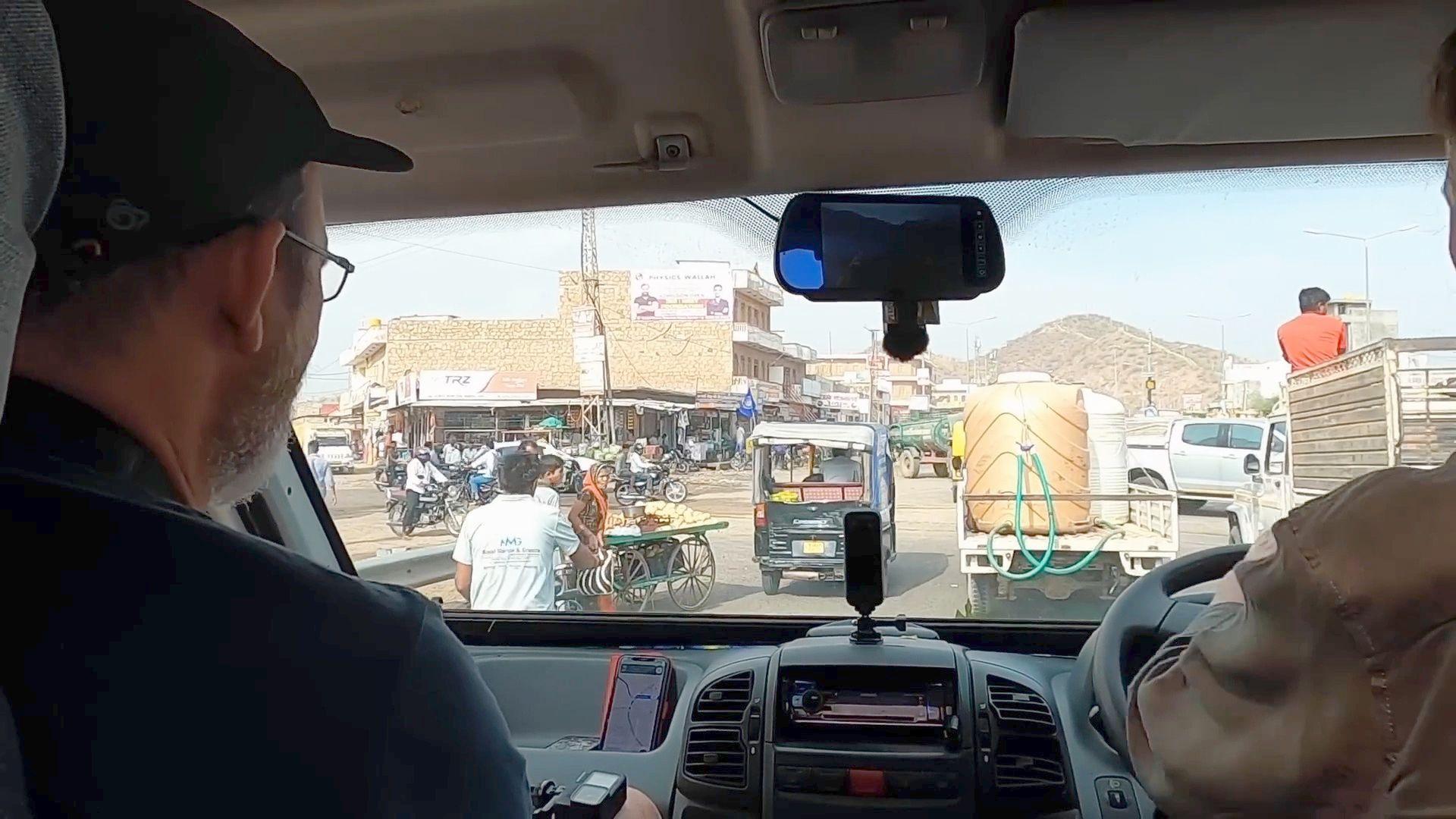 A view from inside a camper van of an Indian road, there is traffic and people ahead and a building to the left side, inside the van a man in a black top is in the passenger seat and someone's arm is just about visible on the steering wheel