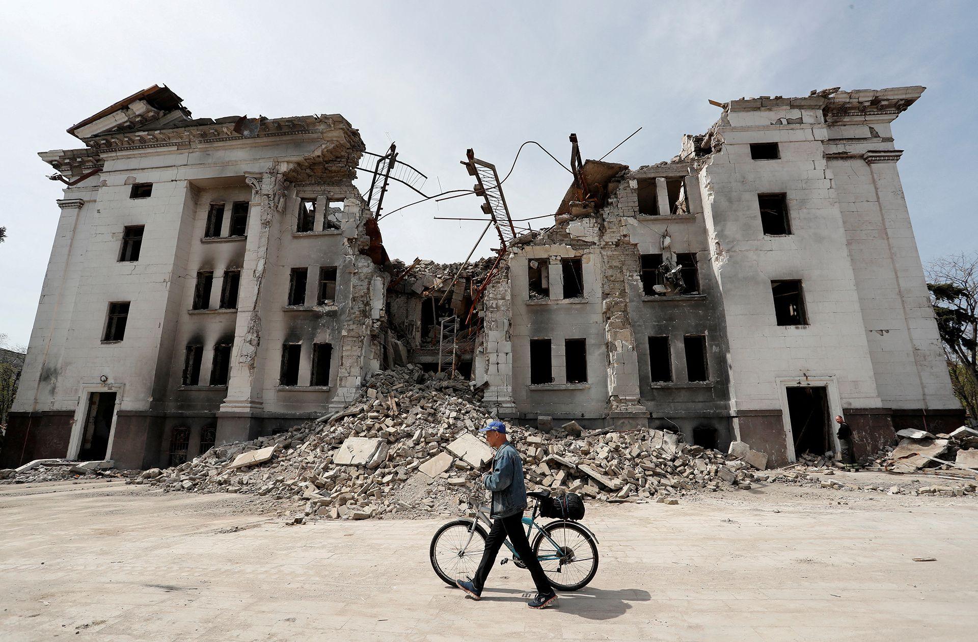 A man walks his bicycle along a street in front of the Mariupol theatre, damaged and half-collapsed surrounded by rubble against a clear blue sky, in Mariupol, eastern Ukraine on 25 April 2022.