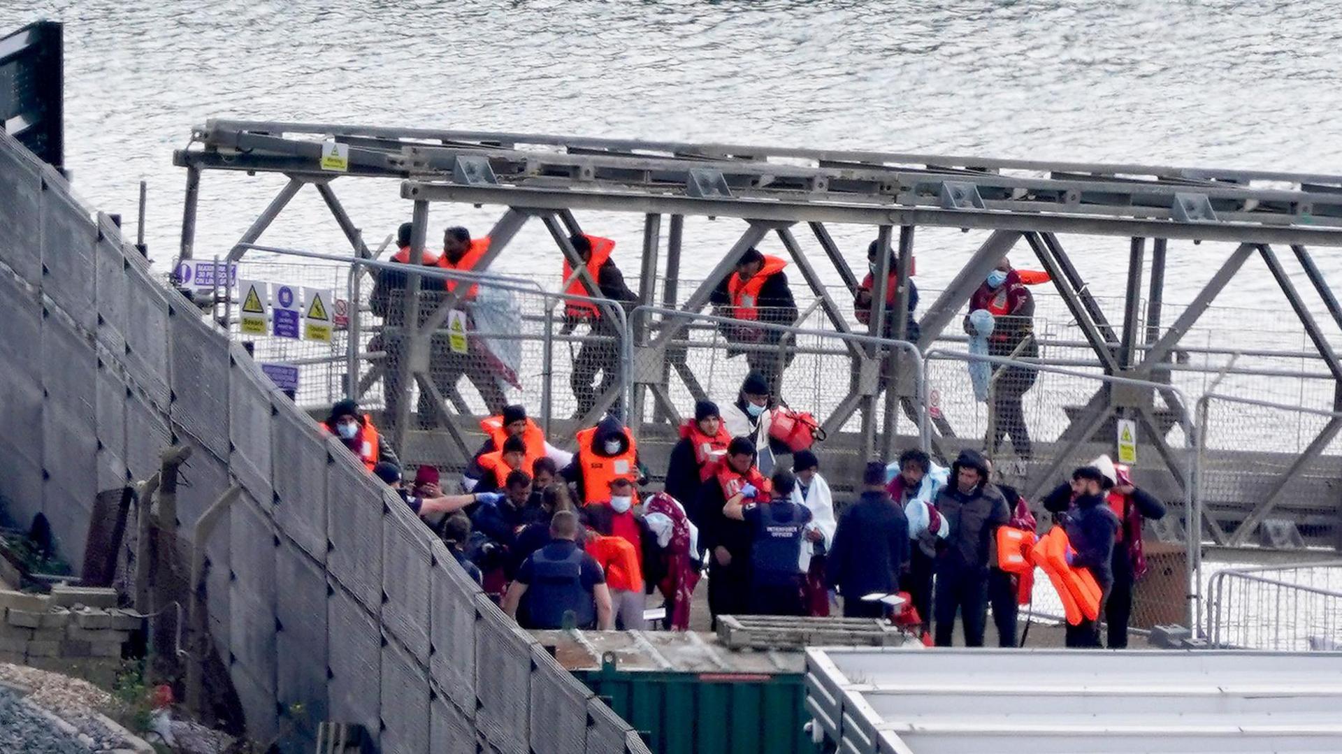 A group of people who have crossed the English Channel in small boats, wearing lifejackets and being led up a gangplank to land at the the port of Dover.