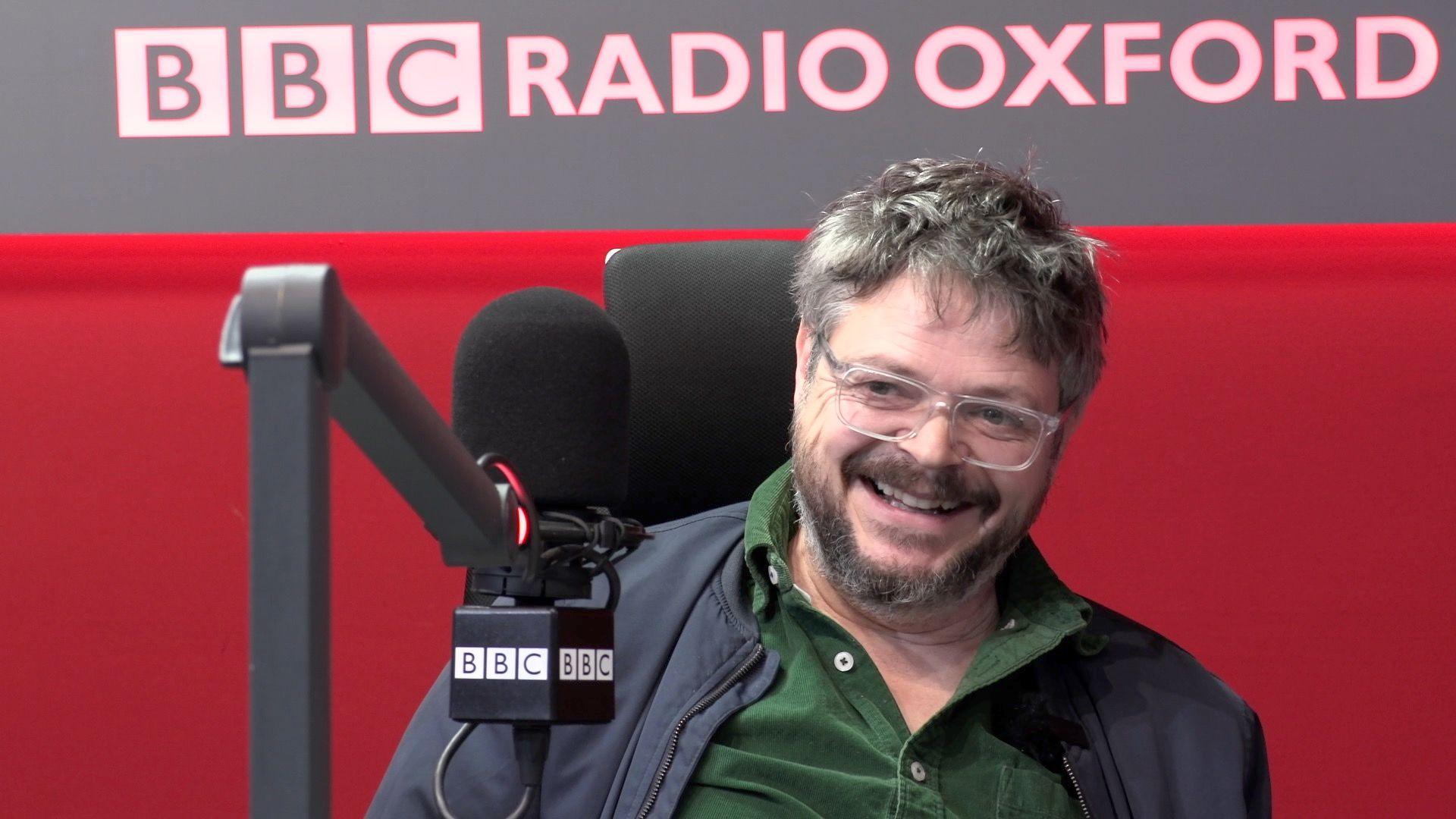 Mick Quinn sitting in front of a microphone and wearing a green, buttoned shirt and a blue jacket. He is smiling as he answers questions. He is wearing clear-rimmed glasses and has greying, short hair