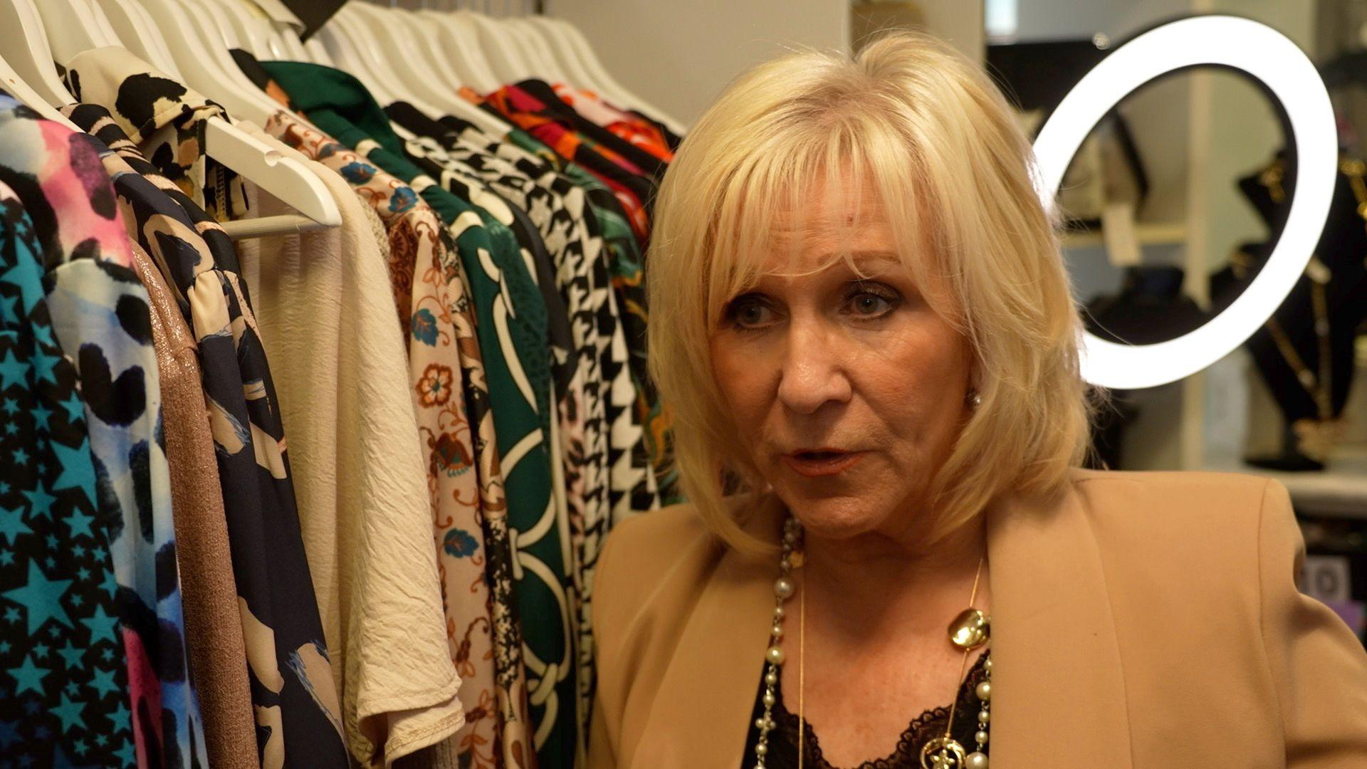 Deirdre Bates is wearing a light brown blazer and is next to a set of clothes hanging from a rail. She is in front of a ring light.