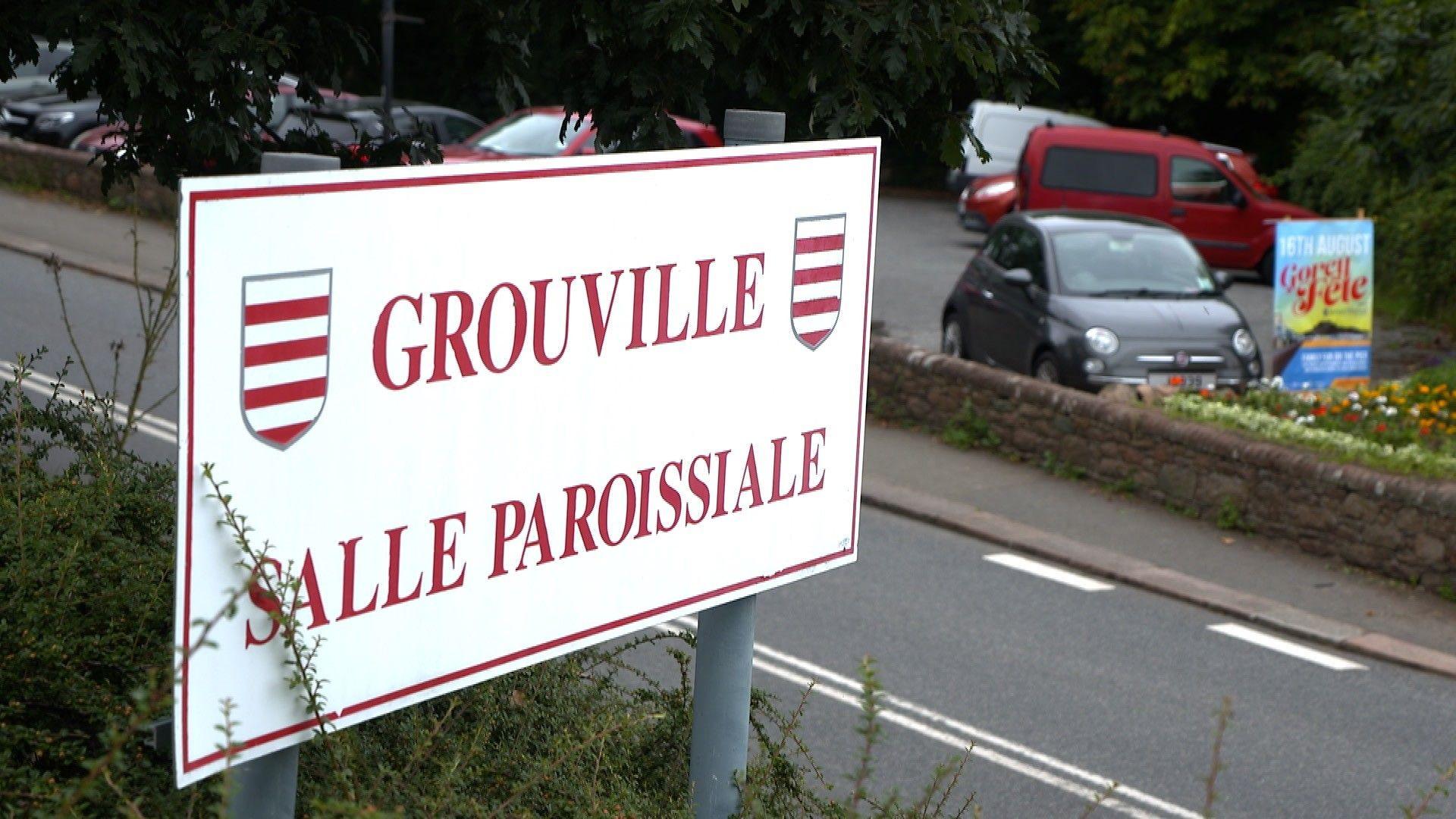 A large white sign with red writing saying Grouville Parish Hall which is next to a road. Behind the road are some parked cars.