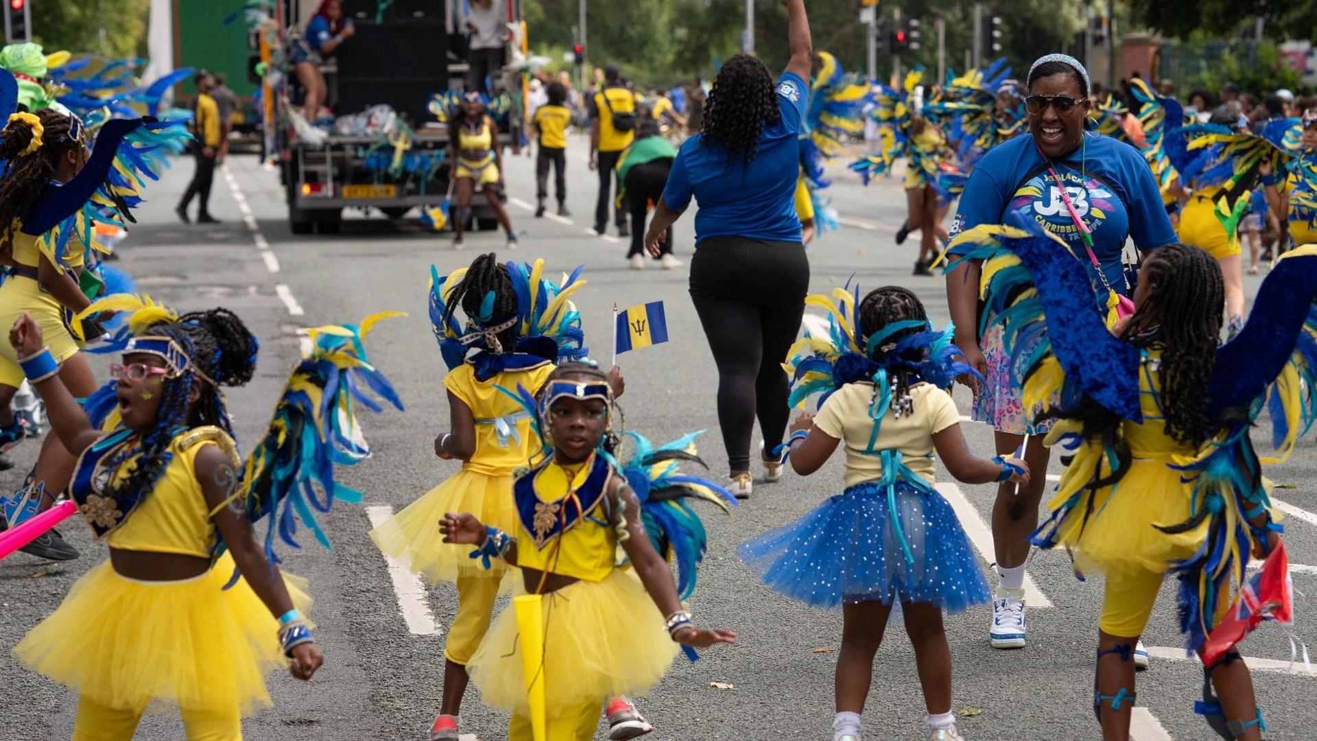 A woman in a blue top grins at five little girls she is leading in a dance during the parade on a road. The girls are wearing yellow dresses with blue wings and trimmings reflecting the colours of the Barbados flag. Other grown-up dancers can be seen in the background behind a lorry in the parade.