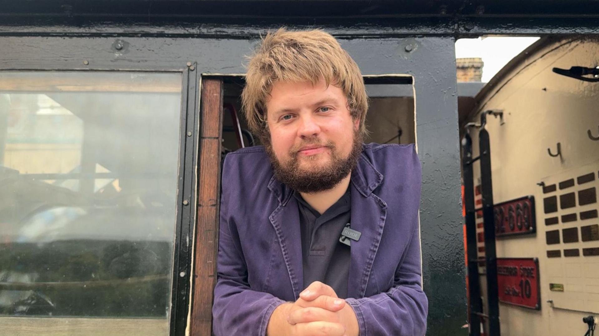 Steam driver Harry Jones leans out of the window of the steam locomotive, facing the camera with his elbows leaning on the ledge and his hands clasped. He is wearing navy blue overalls and has a full brown beard and a head of fair brown hair.