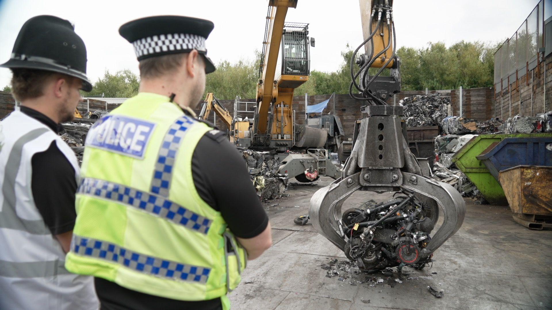 Two officers are stood looking at a pile of crushing e-bikes and e-scooters which are being placed down by a crane. One officer is wearing a yellow and blue police vest and the other is wearing a white hi-vis vest. 