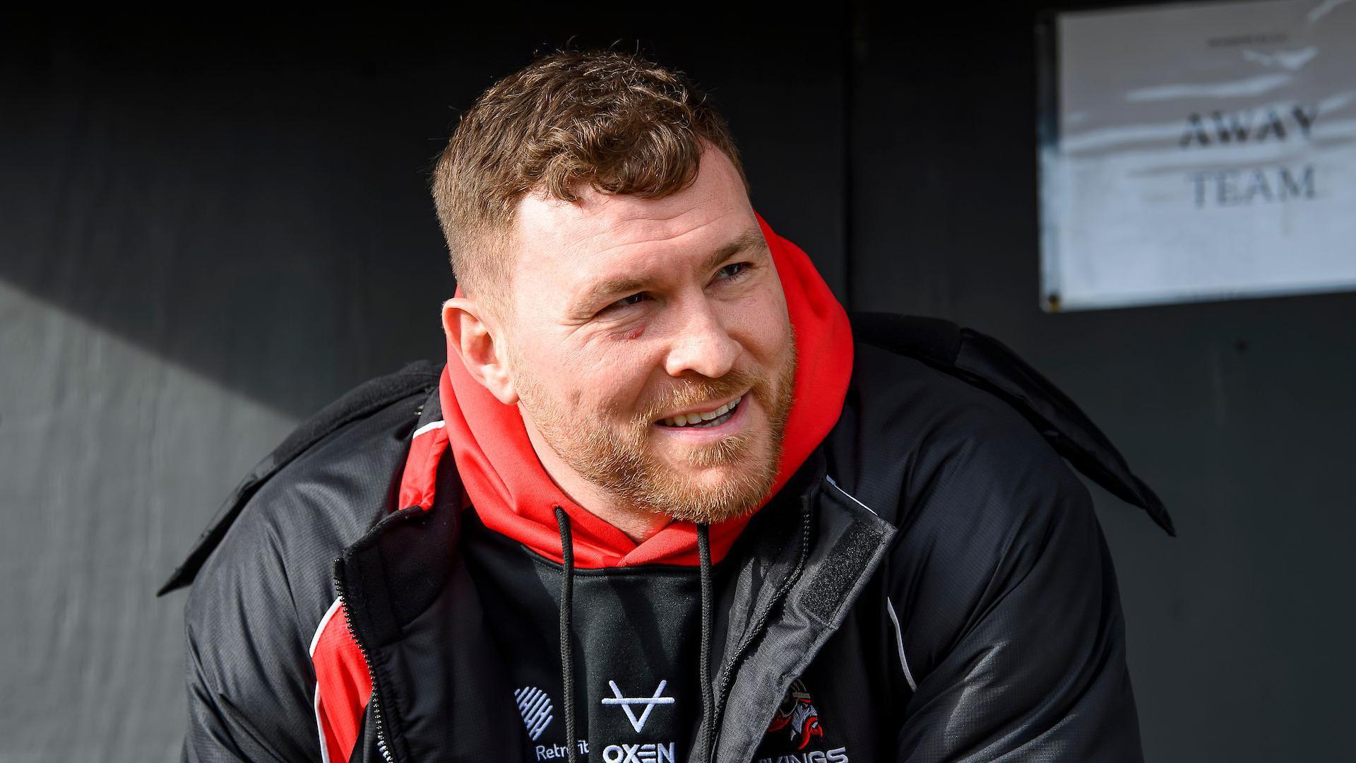 Scott Taylor sat in an away team dugout in his role as Goole Vikings head coach wearing a black rainproof jacket and a red hooded top