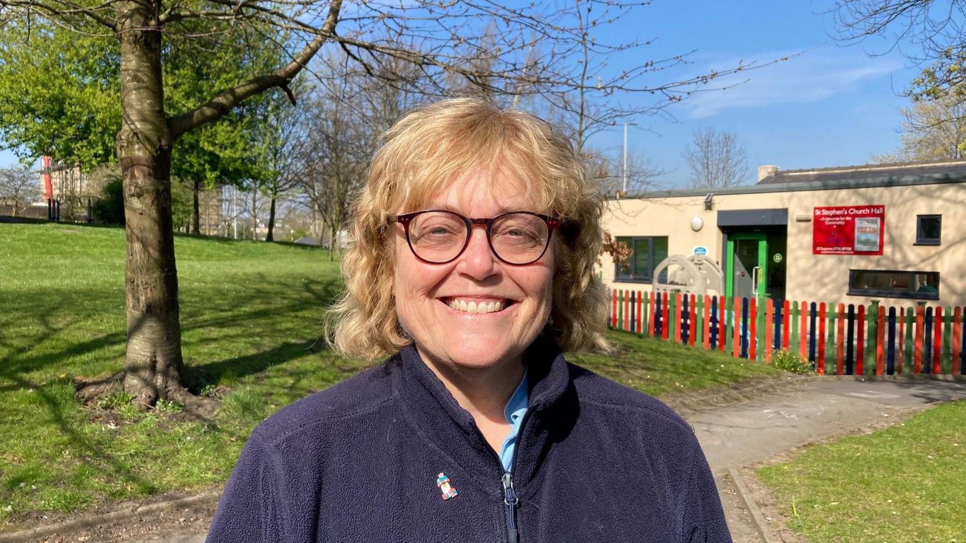 A woman wearing a blue fleece and wearing glasses standing outside. 
