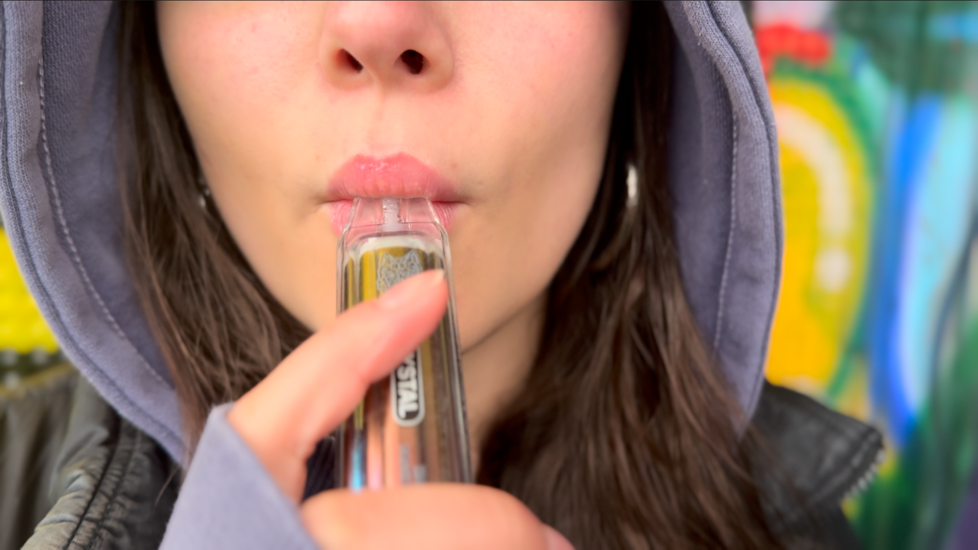 An anonymous teenage girl holds a vape to her lips, just the lower half of her face visible. She is in a purple hoodie, hood up, and black jacket and has long brown hair. She is in a park with colourful graffiti behind her.