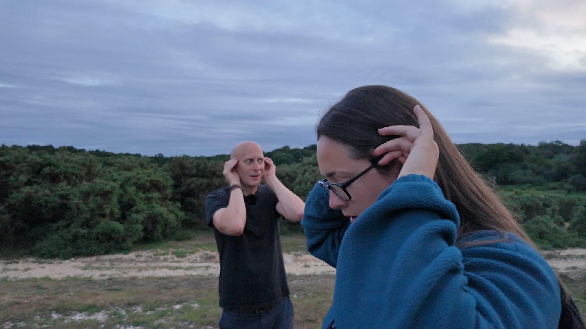 A man and a woman hold their hands up to their ears as they both listen intently for the call of a Nightjar on heathland