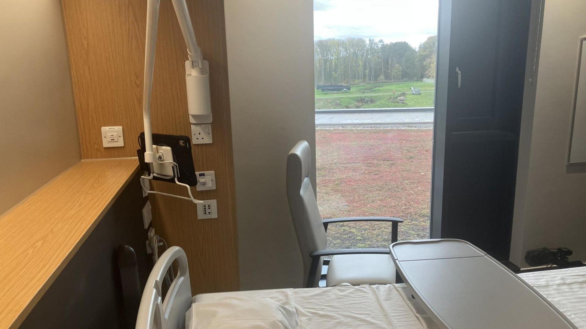 A patient bedroom, kitted out with a brand new bed and other furniture. A big window overlooking parkland that patients can access. 