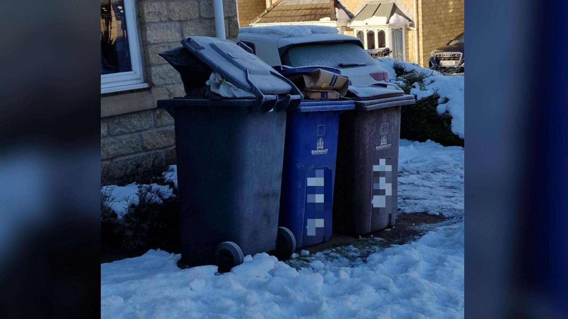 Three bins in a row, two grey an one blue, outside a house with snow on the ground. 