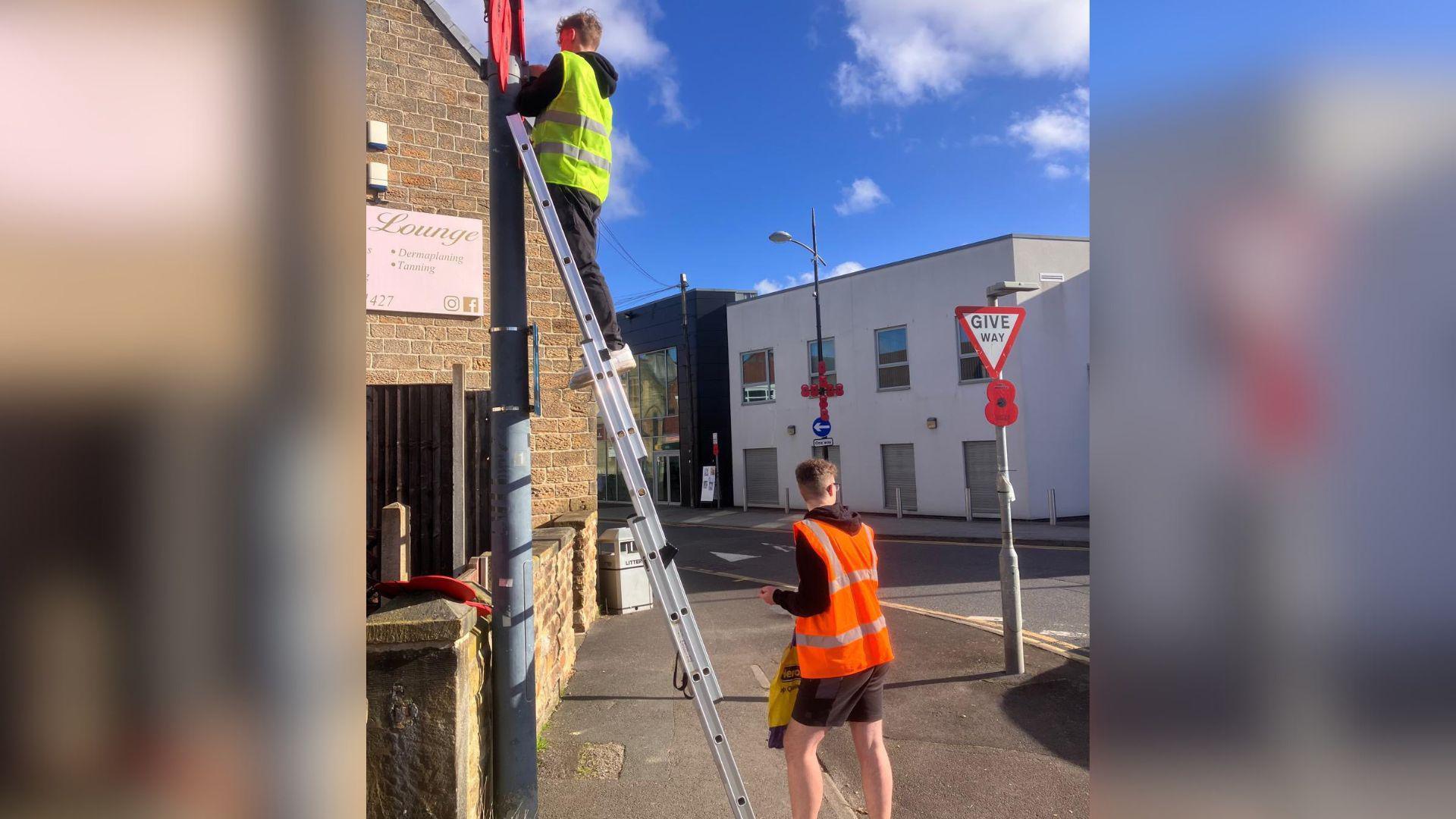 Two men are attaching poppies to a lamppost. One is up a ladder which is leant against the post, while the other stands at the bottom. Both wear high-visibility jackets.