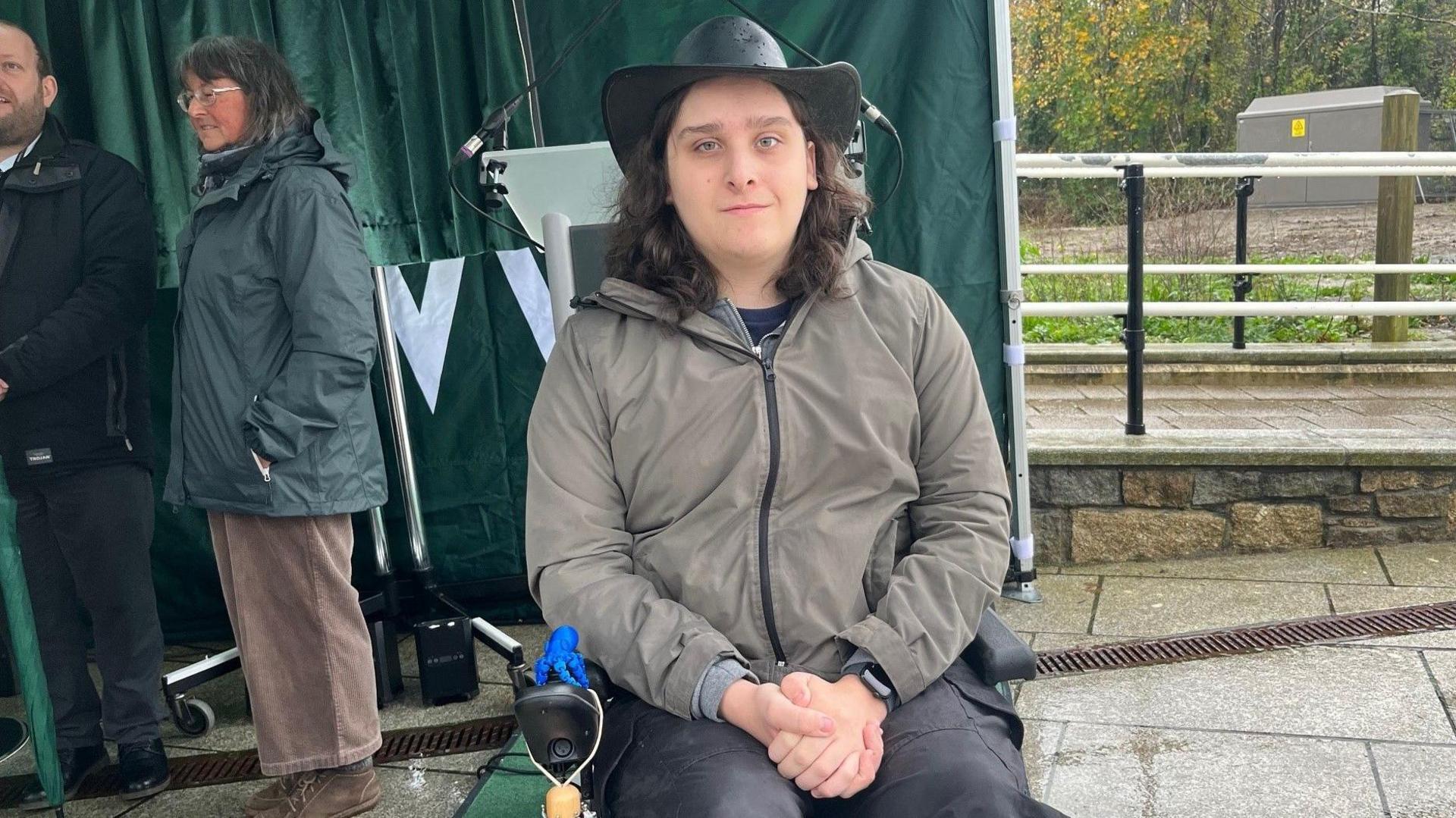 A man in a wheelchair smiles at the camera. He's wearing a black leather rain hat and a grey jacket.