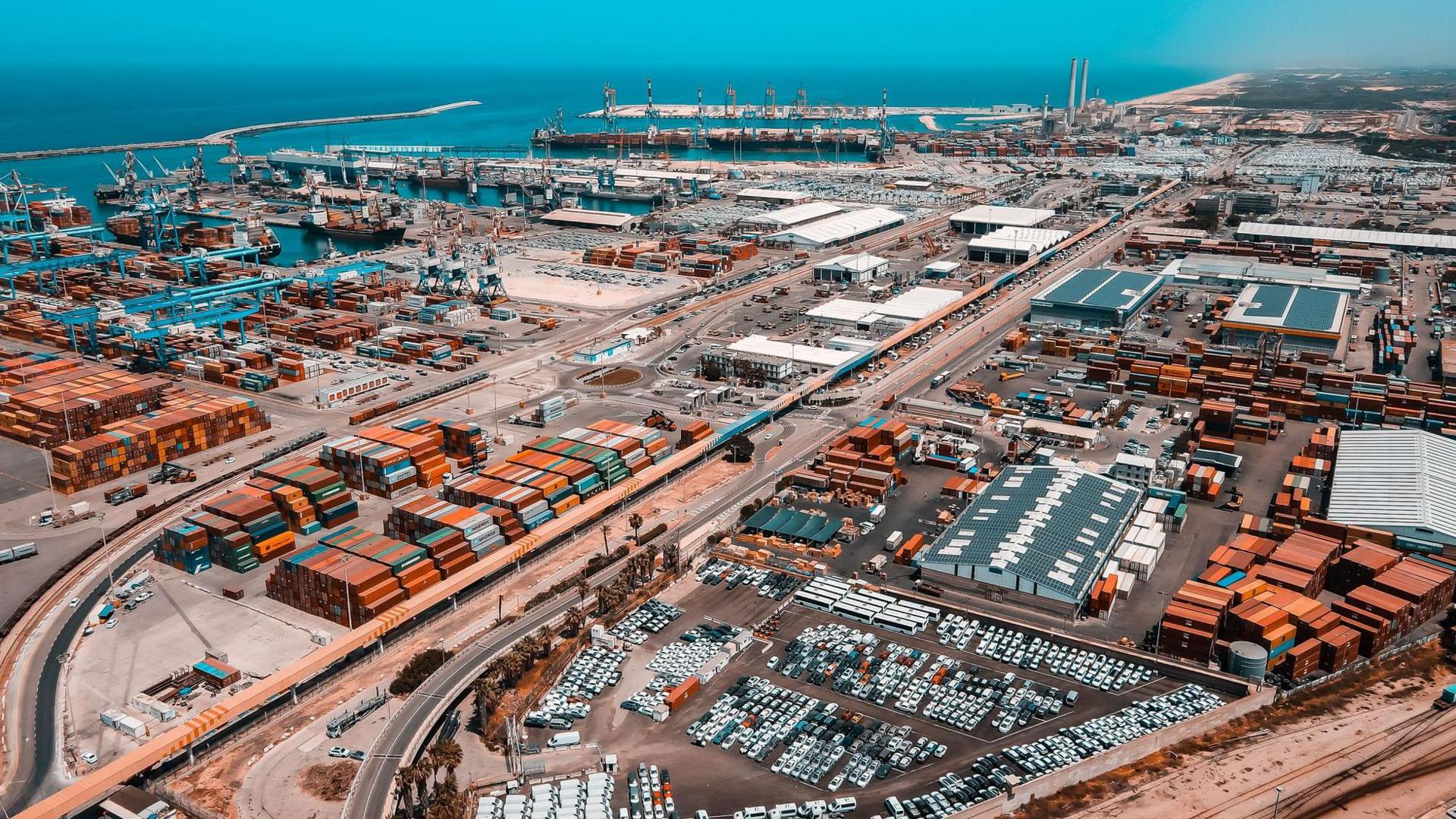 Aerial view of the Port of Ashod. There are multiple orange and green shipping containers in the foreground and a parking area filled with vehicles. There are several warehouses and industrial buildings with white or grey rooftops and there are numerous cranes positioned along the docks. 