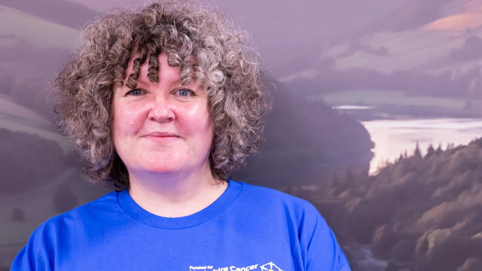 A close-up picture of Karen Nile with shoulder-length curly brown hair. She is smiling and is wearing a blue t-shirt. 