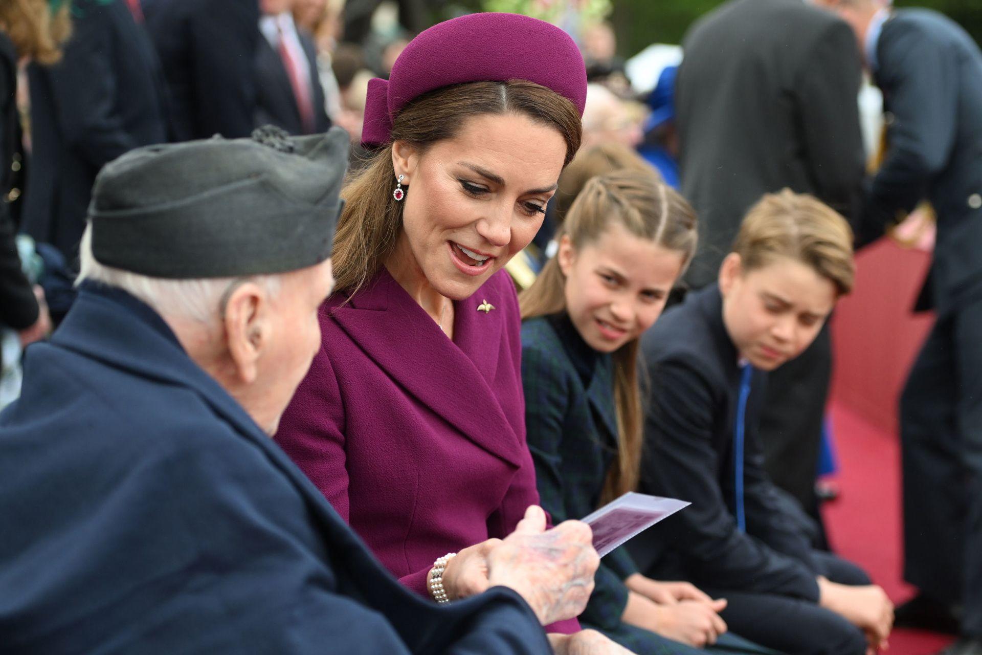 The Princess of Wales looks at some paper while wearing a plum-coloured dress and hat. She's sitting next to a veteran with her children looking on in the background.