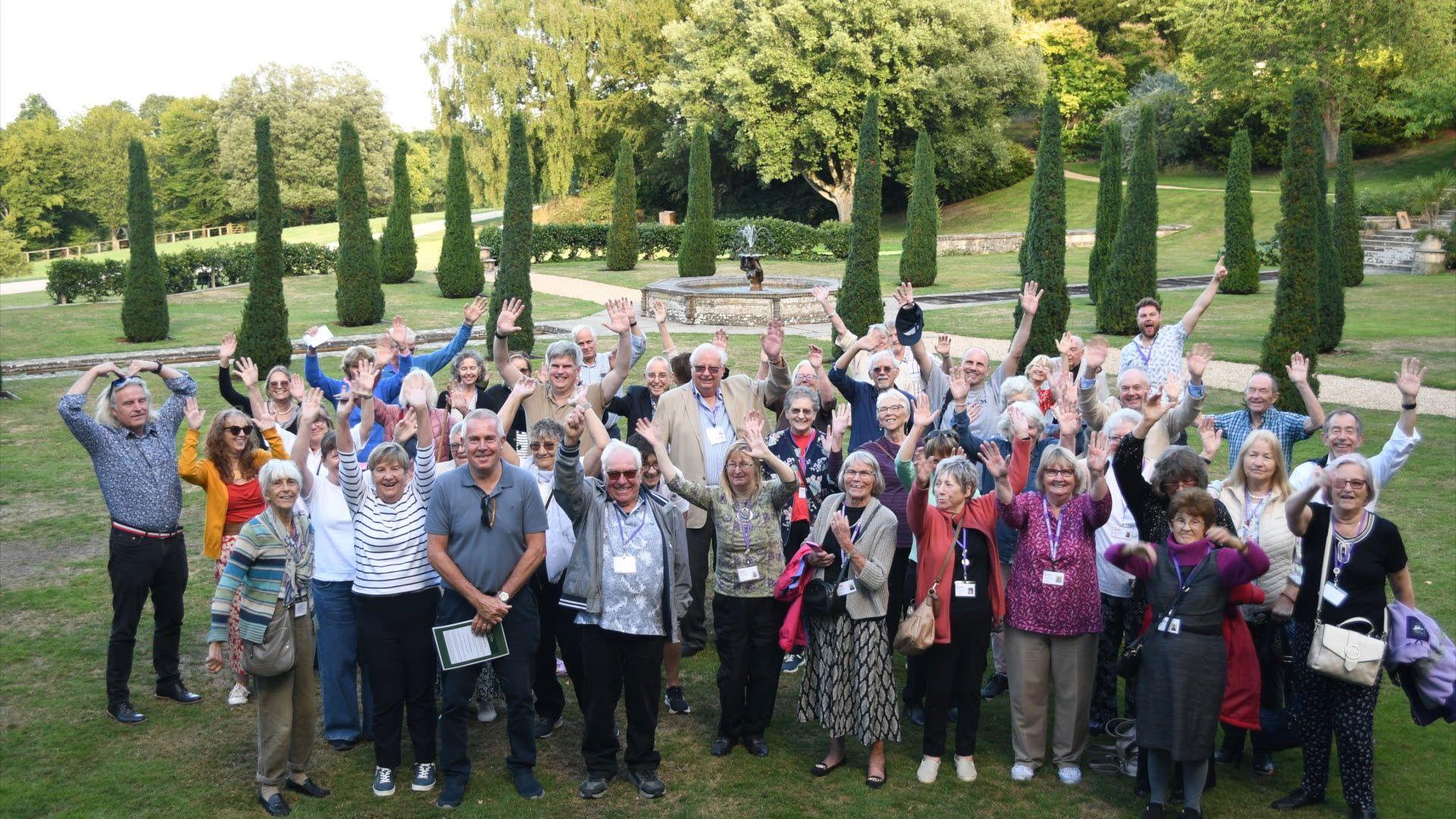 Dozens of people stand in a group smiling and waving at a camera. Many of them have their arms in the air. They are standing in some gardens with about a dozen tall, thin pointed hedges, large trees and a manicured lawn in the background.