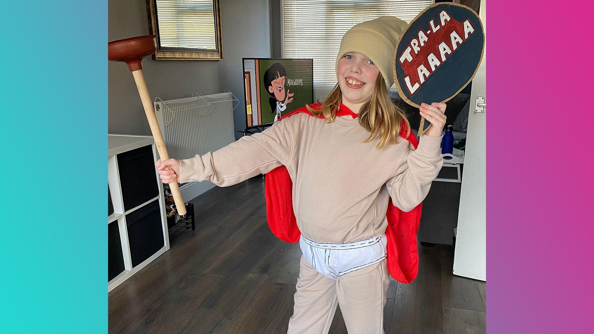 Tango is wearing a beige tracksuit with white underpants over the top. They are wearing a red cap and holding a plunger in one hand and a home made sign that says 'Tra-la-laaaaa' in the other 