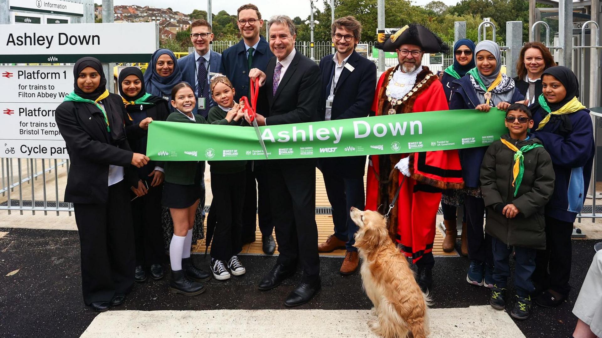 A group of people, including West of England mayor Dan Norris and local MP Darren Jones, stand in front of a green ribbon at the opening of the new Ashley Down Station in Bristol. Local schoolchildren are also pictured, as is the city's lord mayor and Dan Norris's dog