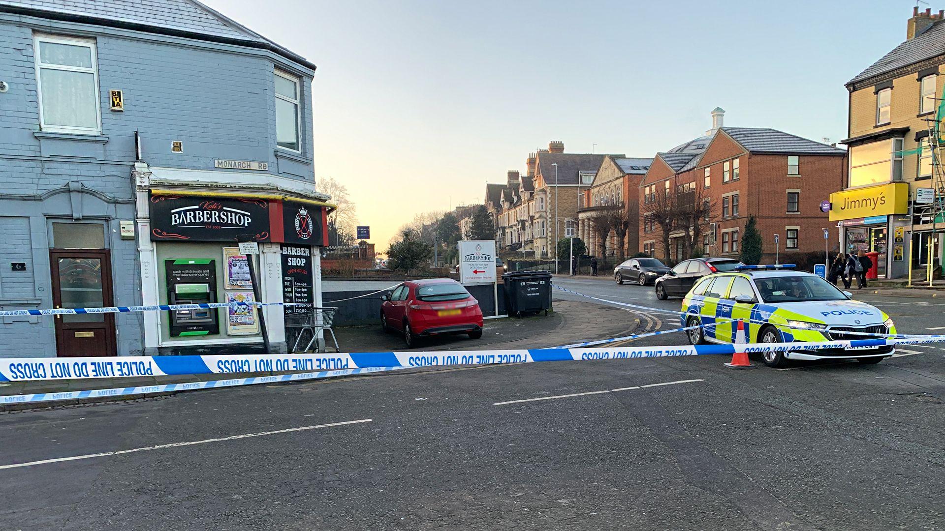 A police cordon on Monarch Road, at the junction with Kingsthorpe Road, in Northampton. You can see police tape across the junction close to a barbers' shop. A red car is within the cordon. A police car is also visible.