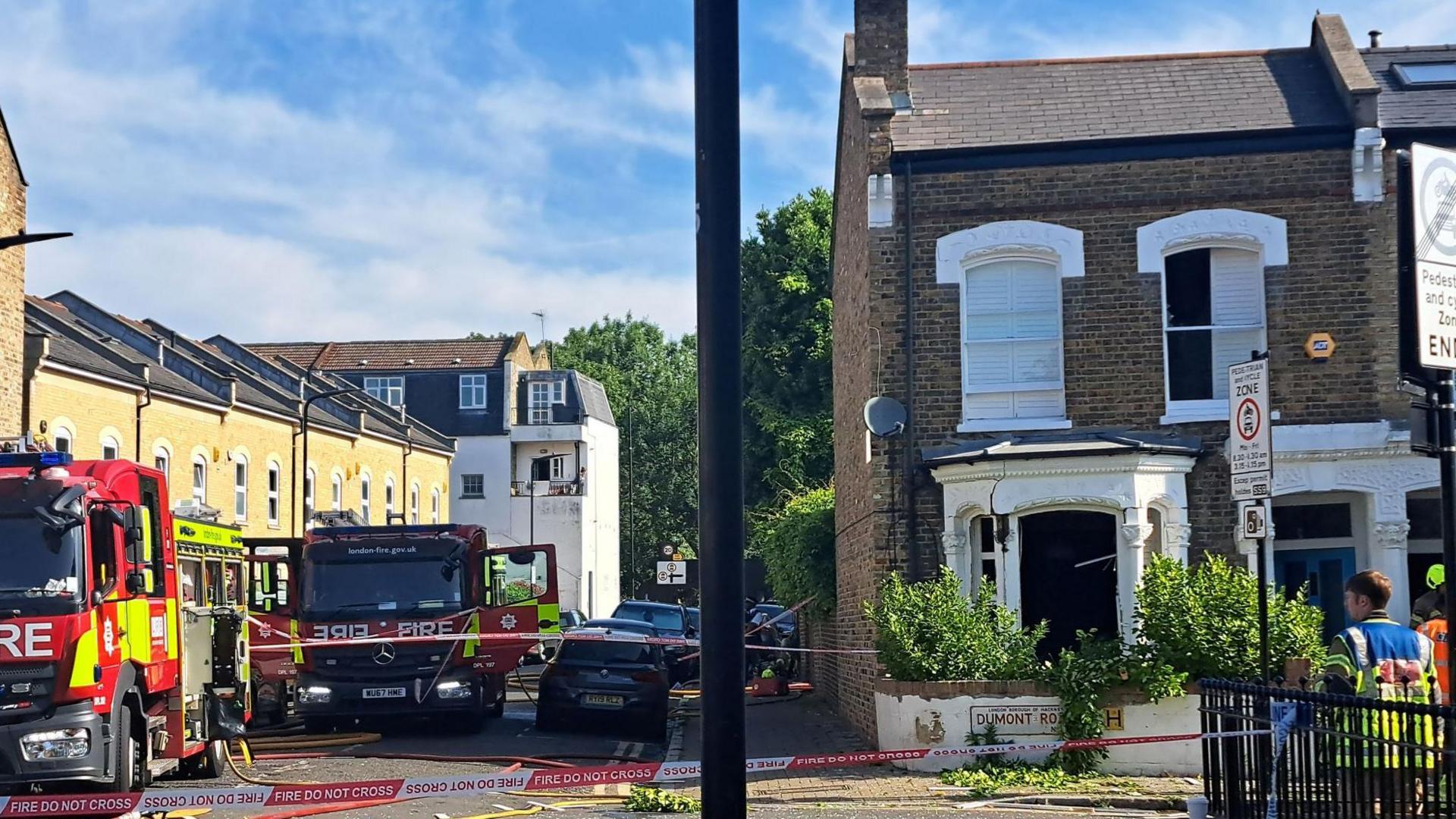 A house with a blown out window on a residential street, with fire engines parked along the road