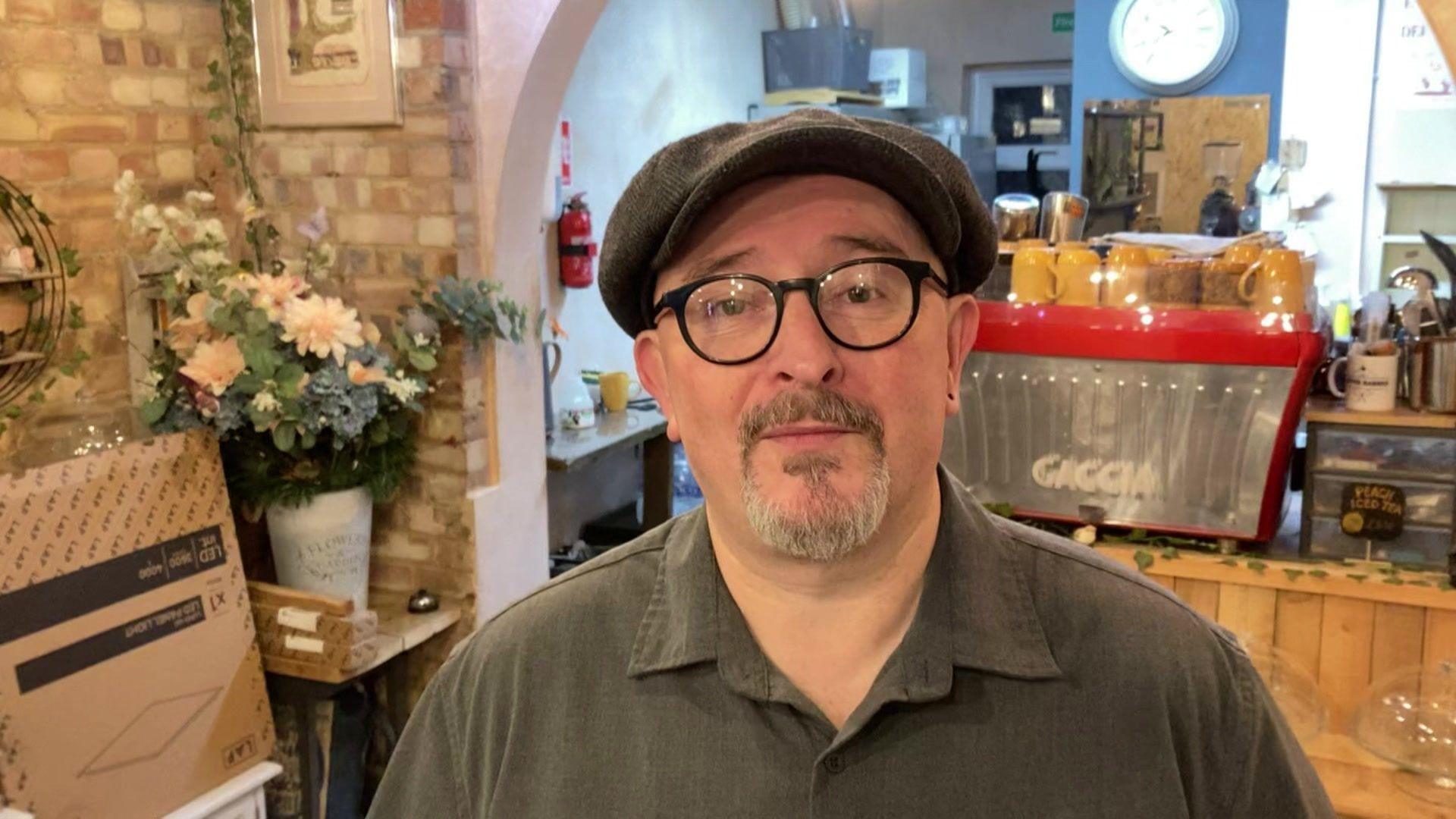 Daren Wayland stands in his coffee/barber shop and faces the camera. He wears a dark green flat cap, with black round glasses and a green shirt. He has a piercing in his left ear. The coffee shop can be seen behind him.