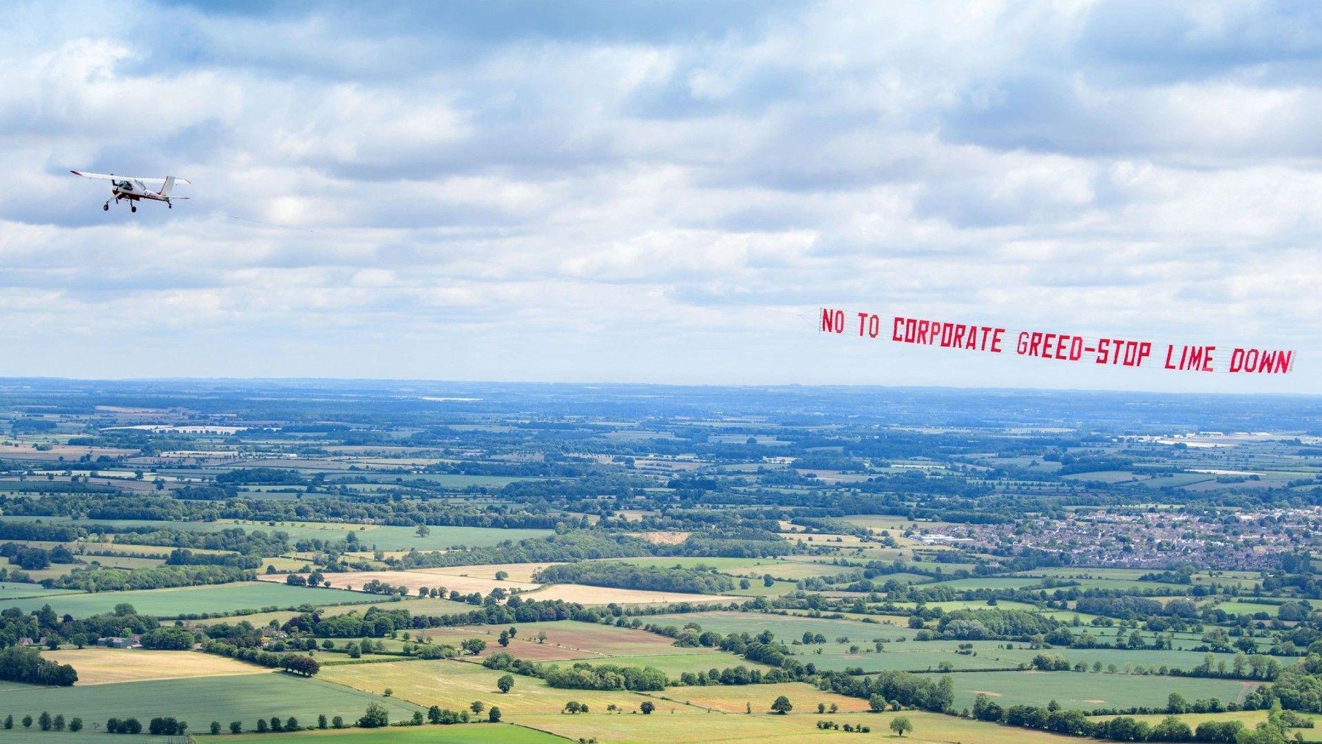 A small plane is in the sky over some countryside, behind it is a banner which reads 'No to Corporate Greed - Stop Lime Down.'