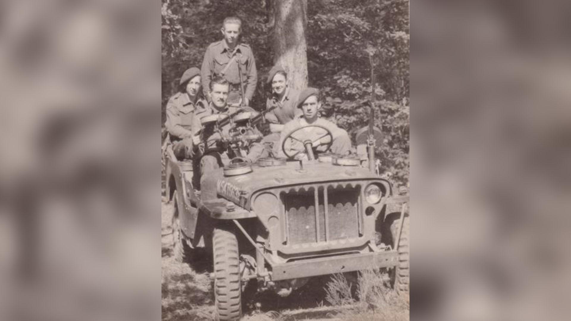 Jim Almond is sitting behind a Vickers machine gun mounting in the front of a Land Rover. Four other SAS men are in the vehicle. They are pictured in a clearing in a forest, with dense woodland in the background.