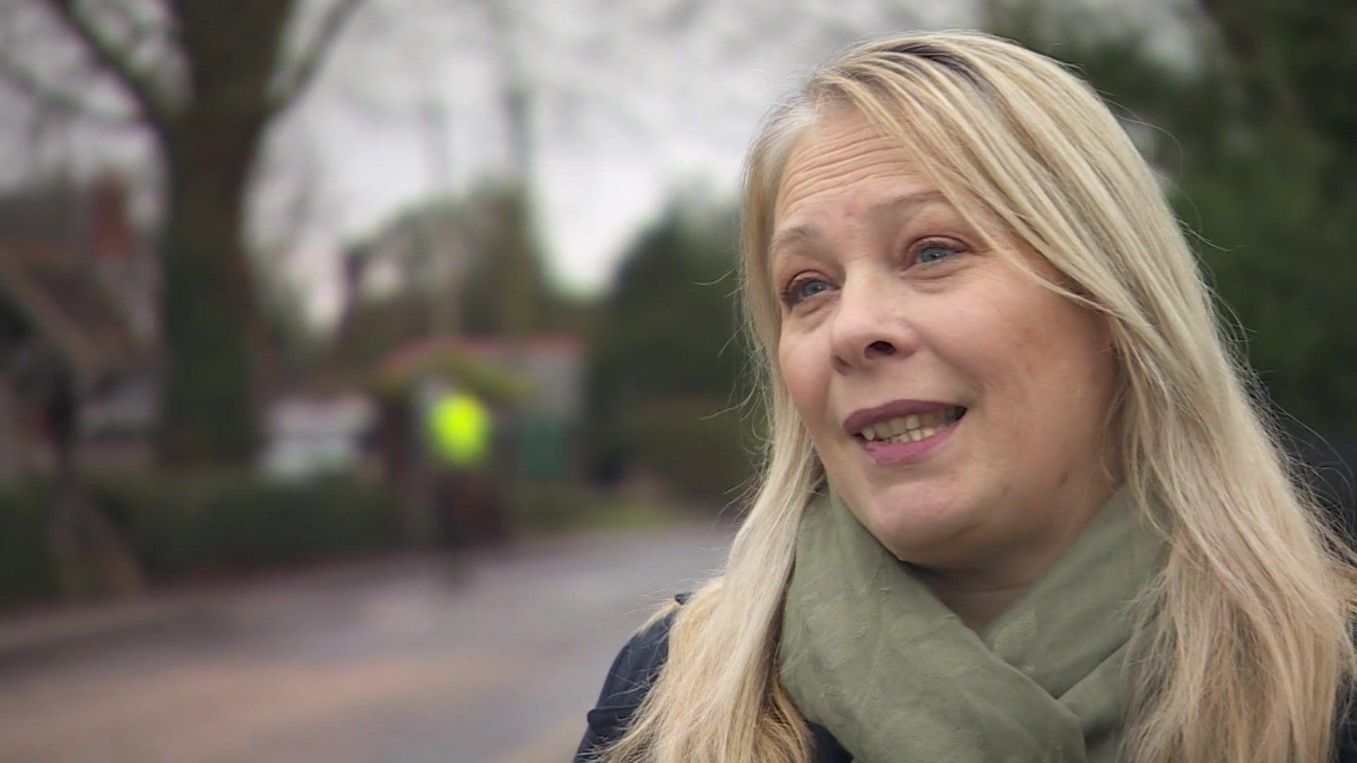 Hannah Colls has long blonde hair and is wearing a green scarf and blue coat. Behind her out of focus is a road.