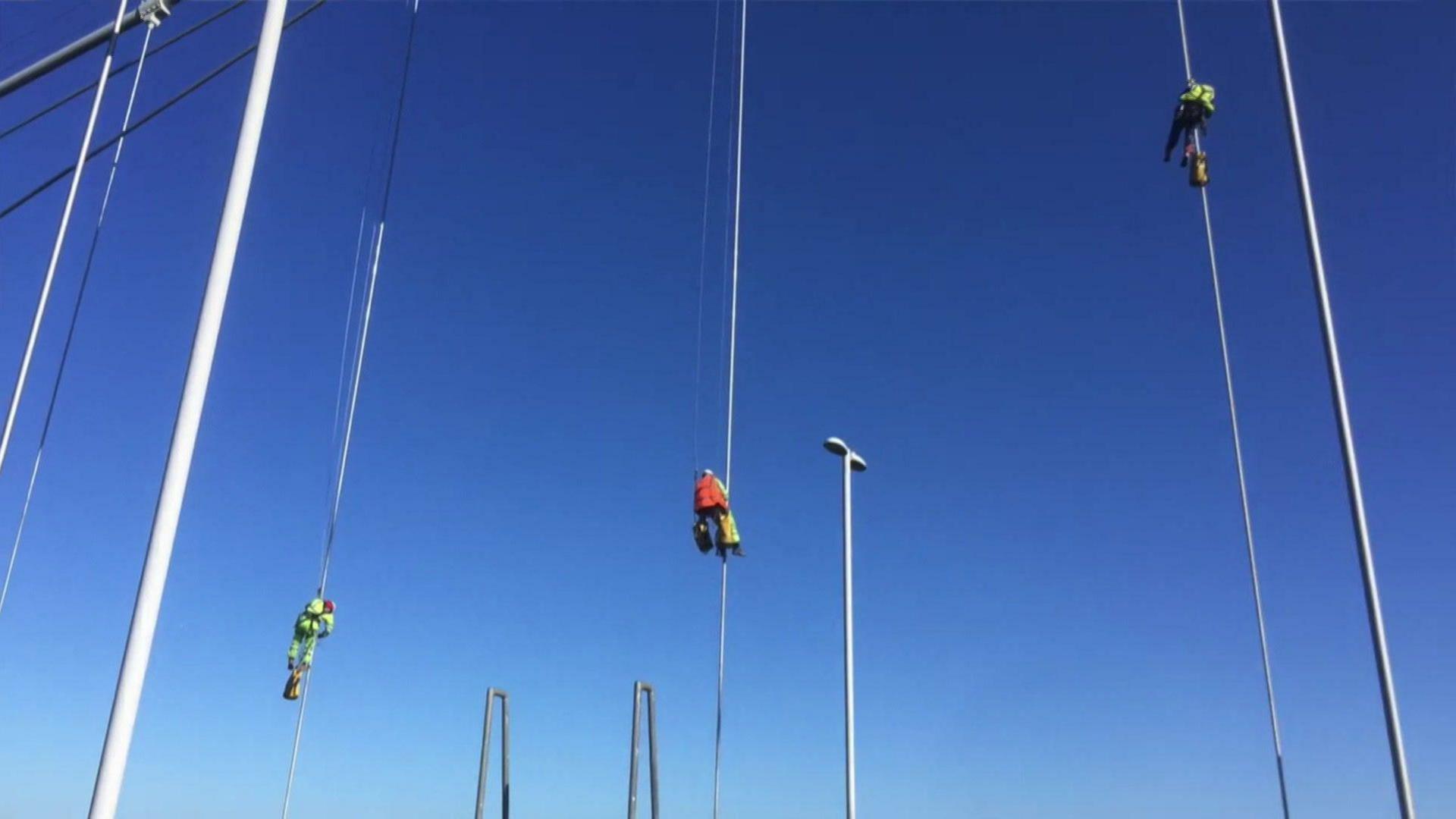 Three men wearing high-vis overalls and hard hats suspended on the cables of a suspension bridge. The sky in the background is bright blue.