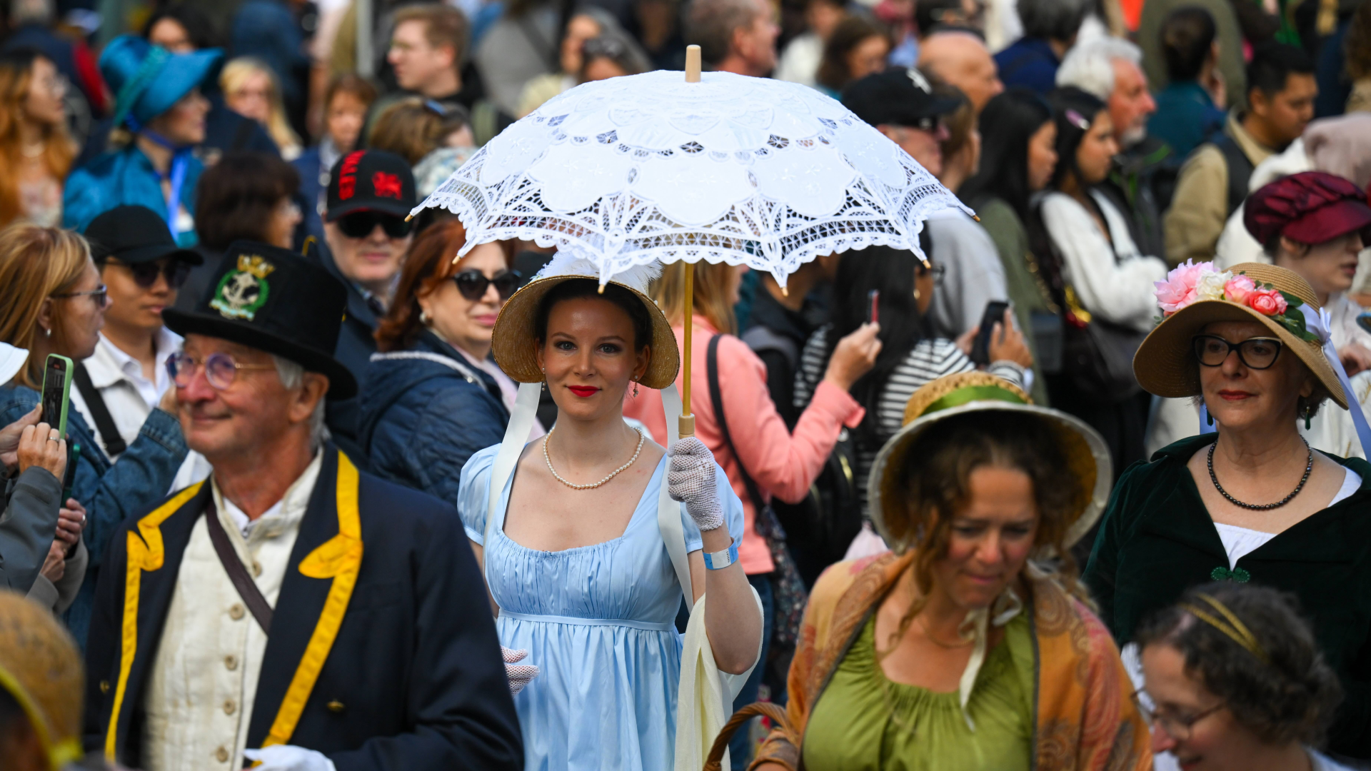 A large crowd of people dressed in regency costumes for the Jane Austen Festival. The image is focused on a woman wearing a blue dress, a bonnet and carrying a white lace parasol. 