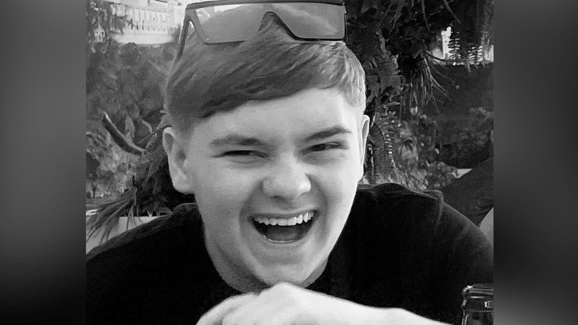 A black and white picture of the teenager who is laughing. He wears sunglasses on the top of his head, trees and plants can be seen in the background.