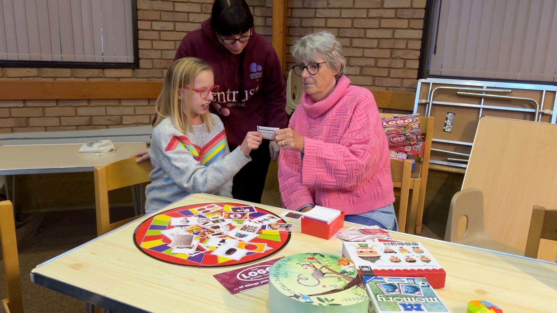 A worker at Entrust Care Partnership takes part in a game with a child and adult, as they sit at a table, which is laden with board games.