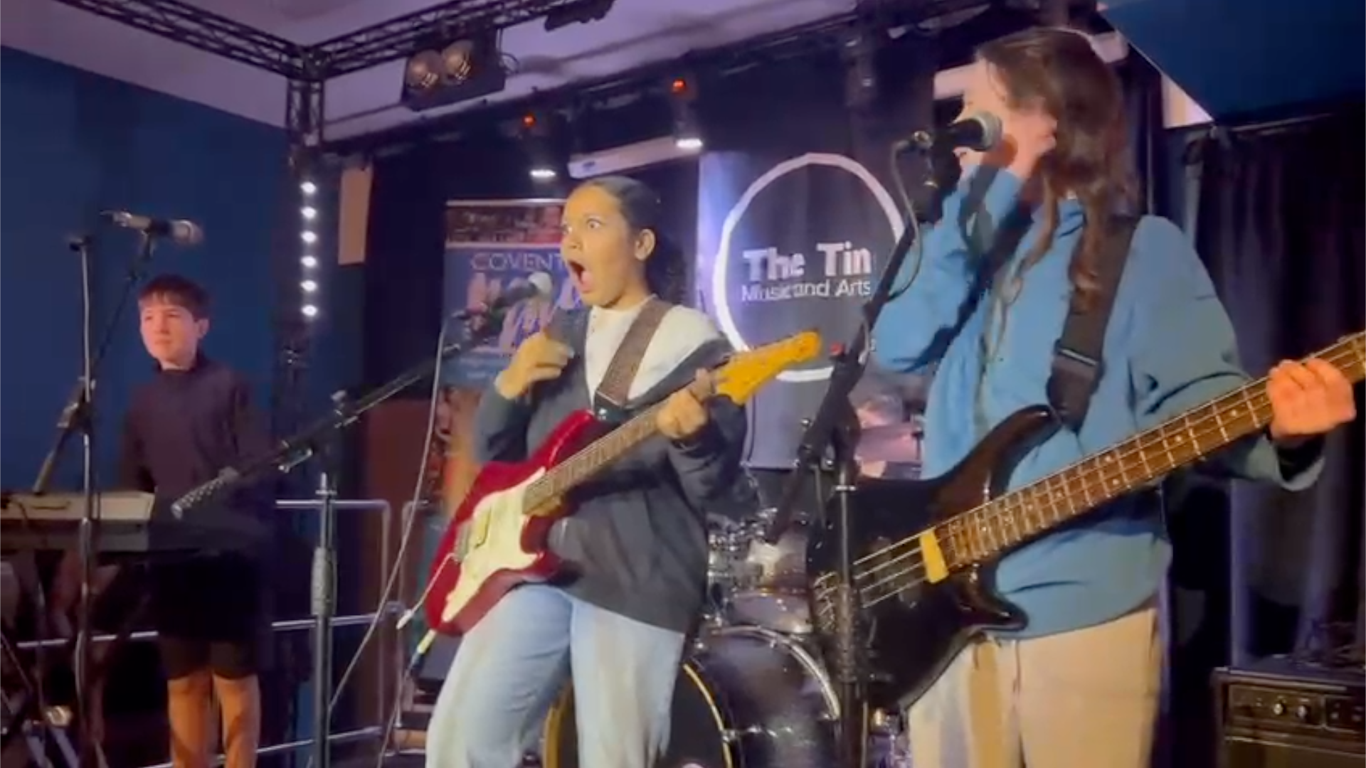 A student holding a red electric guitar with a shocked expression on her face, as she stands behind a mic, with a bandmate on keyboards to her right and the bass player to her left.