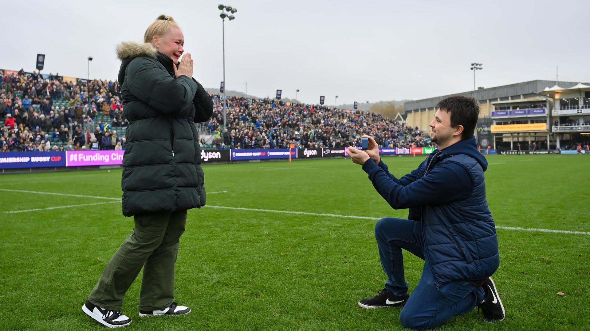 A man is down on one knee as he proposes at The Rec in front of a crowd. His partner - a woman with blonde hair - is holding her hands together and crying.