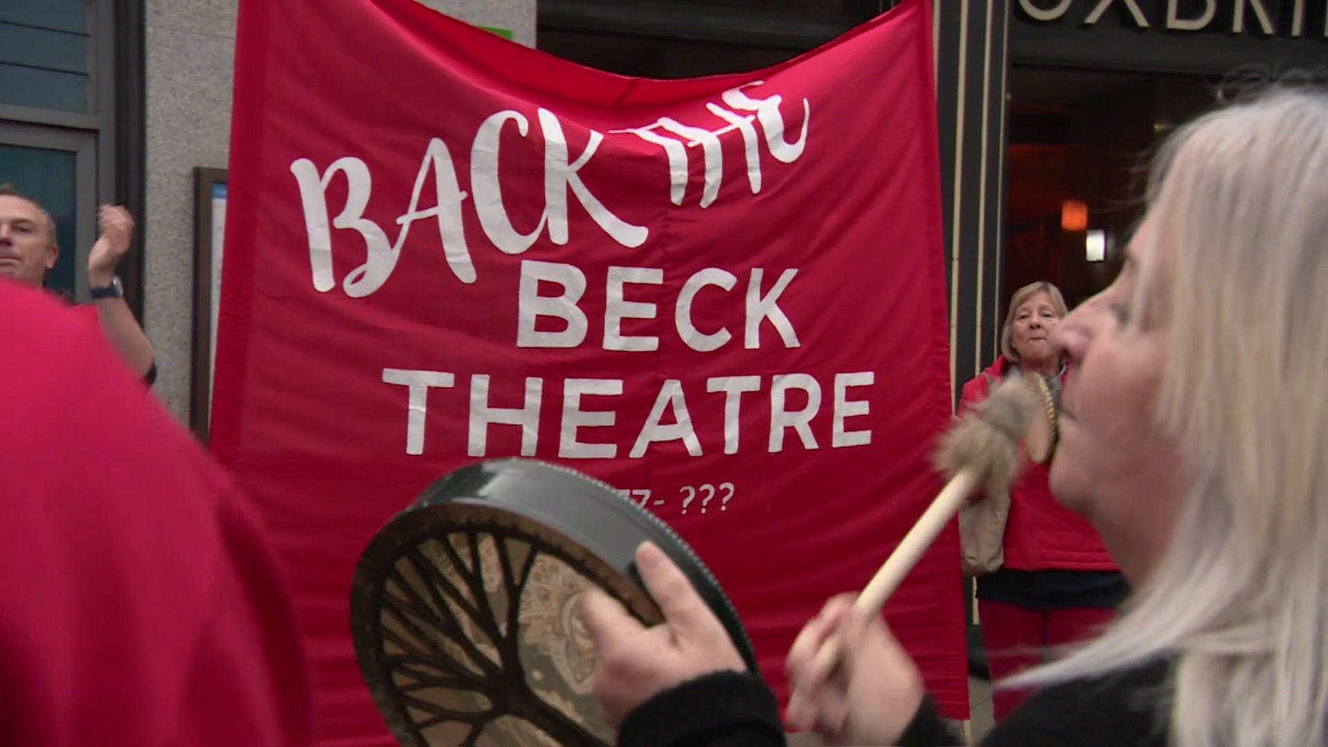 A woman bangs an instrument as protesters clap and hold a red banner sign saying "Back the Beck Theatre".