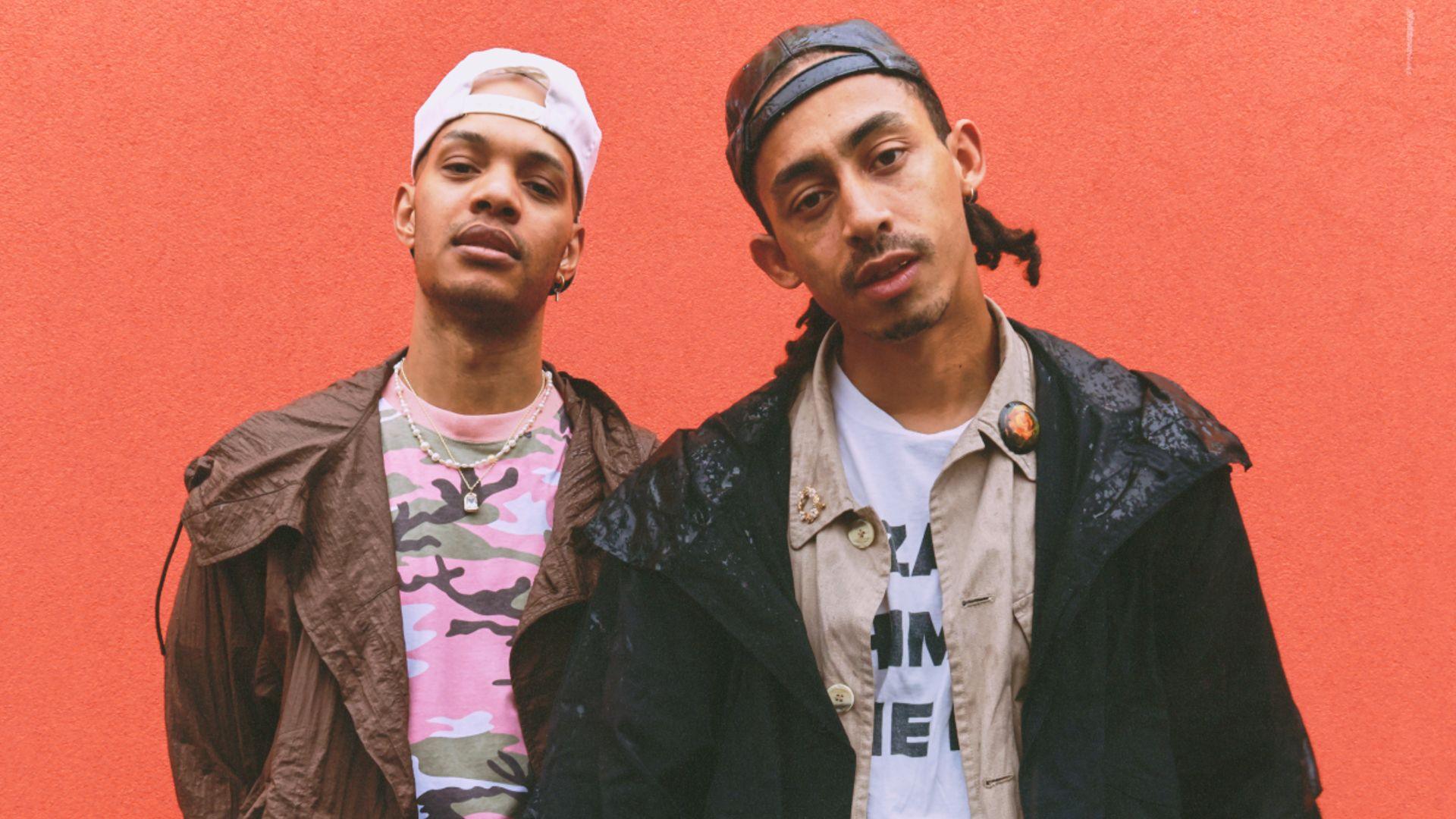 Harley Alexander-Sule and Jordan Stephens, aka Rizzle Kicks, look into the camera. Harley is wearing a pink camouflage t-shirt with a brown coat and backwards white baseball cap. Jordan is wearing a white t-shirt with a beige shirt and black coat over it, with a black baseball cap, also worn backwards. They are standing in front of a tomato-red background.