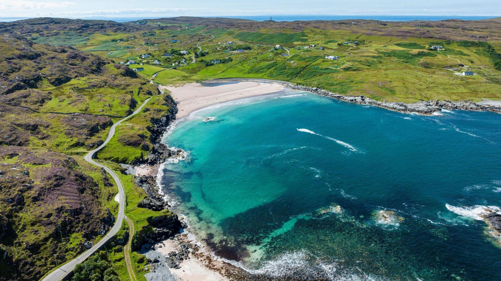 An aerial view of beautiful blue sea waters and the sandy beach, which is surrounded by a green and rocky landscape.