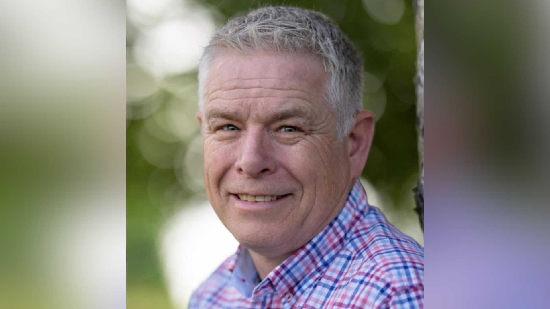 Stuart Williams looks to his left at the camera and smiles. He has short grey hair. He wears a blue and red chequered shirt. 