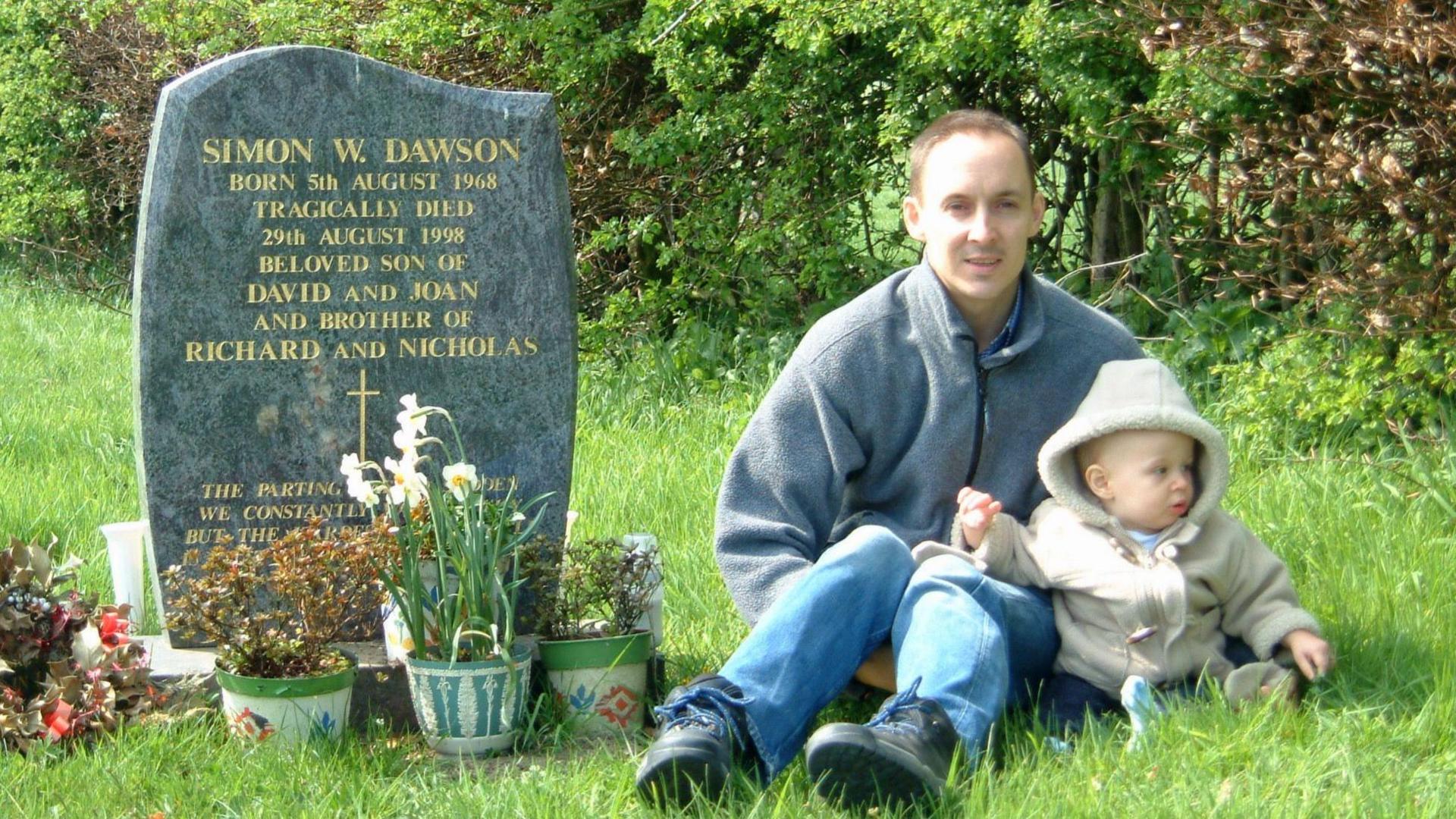A man holding a young child sat next to a grave. The grave has a black stone with gold writing reading: "SIMON W. DAWSON".