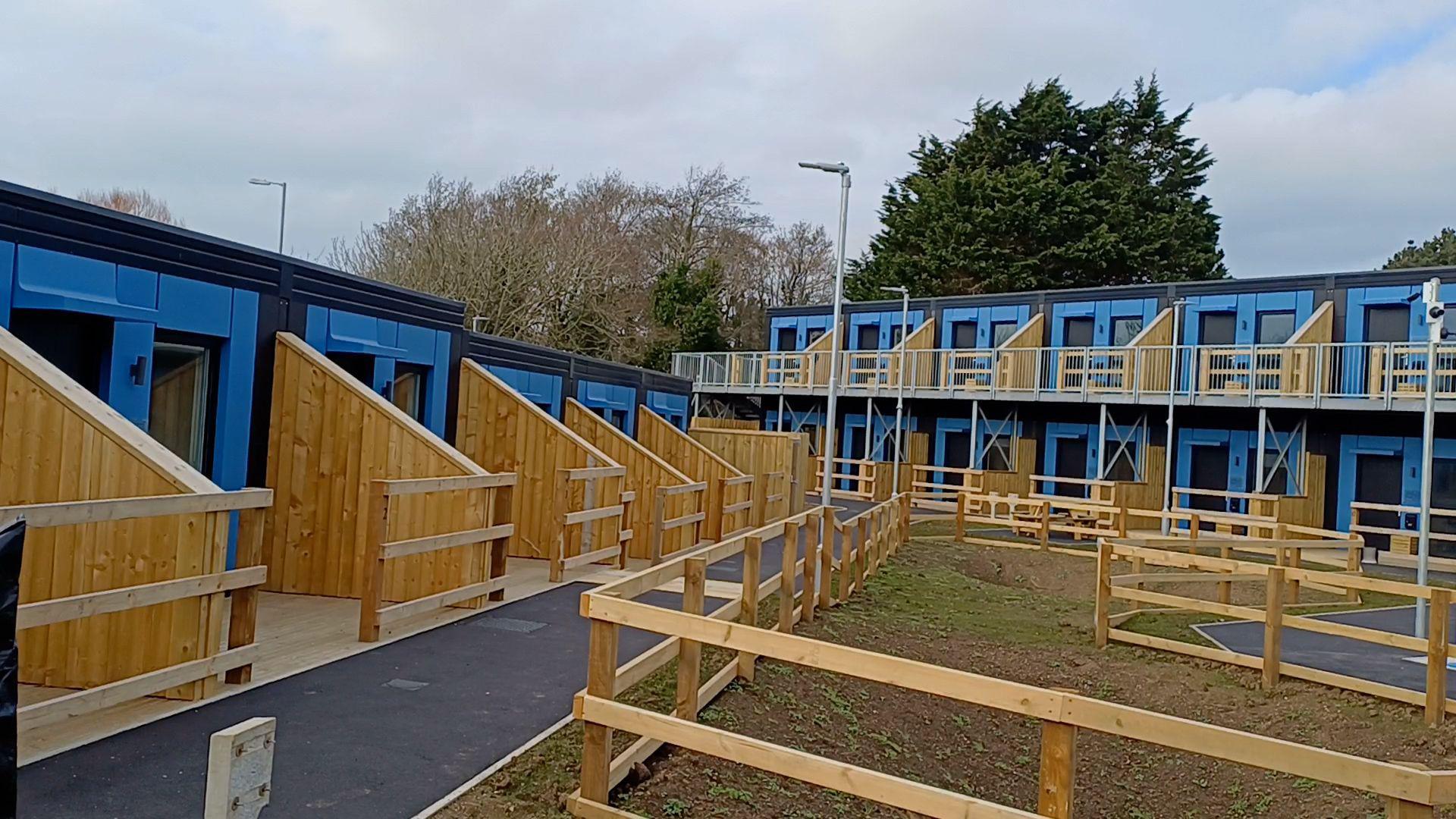 Exterior of the terraced units. A row of four, on the left hand-side, are separate by wooden panels. Additional units, on two storeys, are opposite. They are also separated by wooden panels. The units are blue and black.