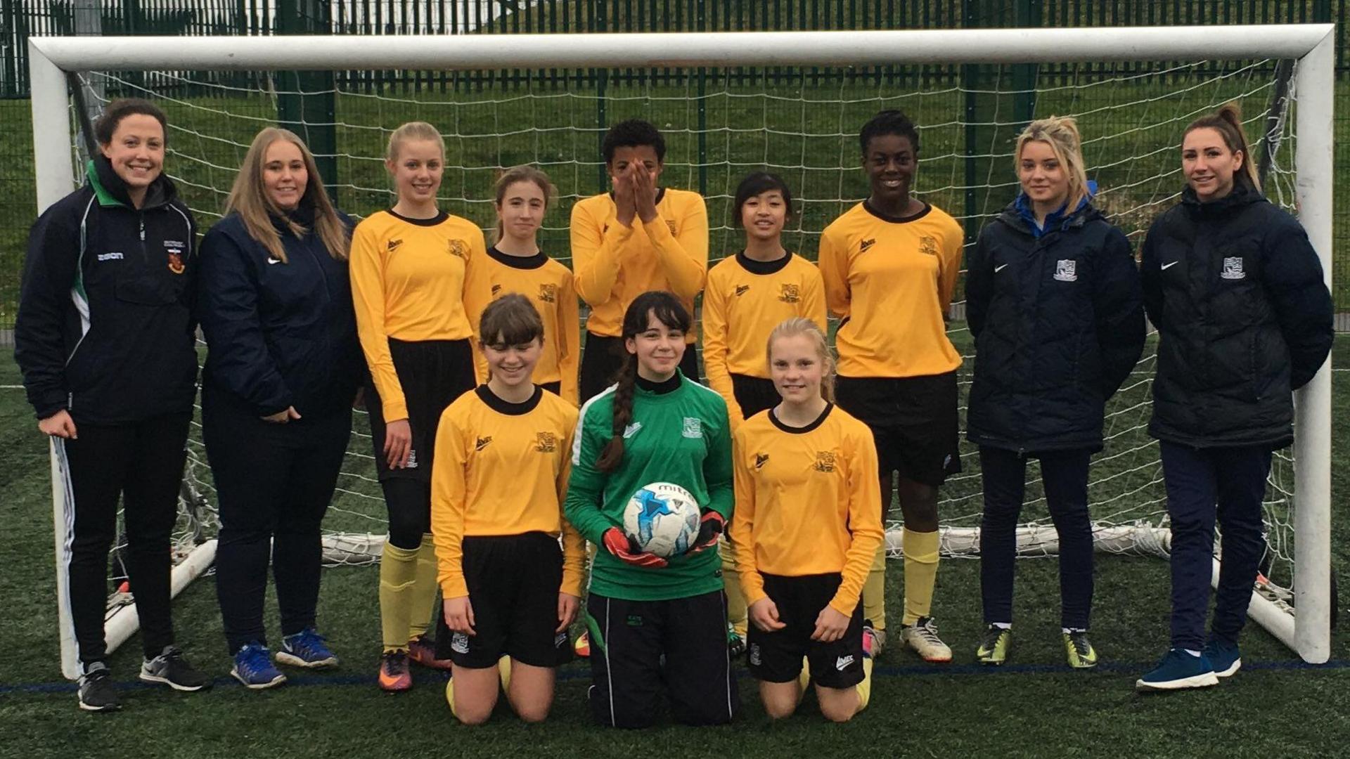 Eight girls in yellow football tops with four coaches standing either side of them, all of them are posing in front of a goal