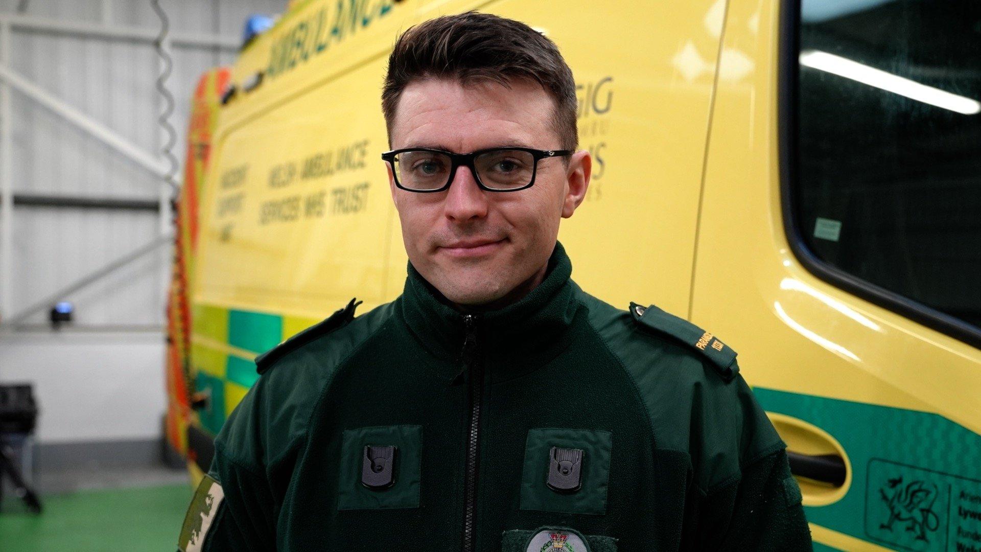 Gareth has short, dark hear and is wearing glasses. He is wearing his green Welsh Ambulance Service uniform and is stood next to one of the large vans containing their specialist kit.