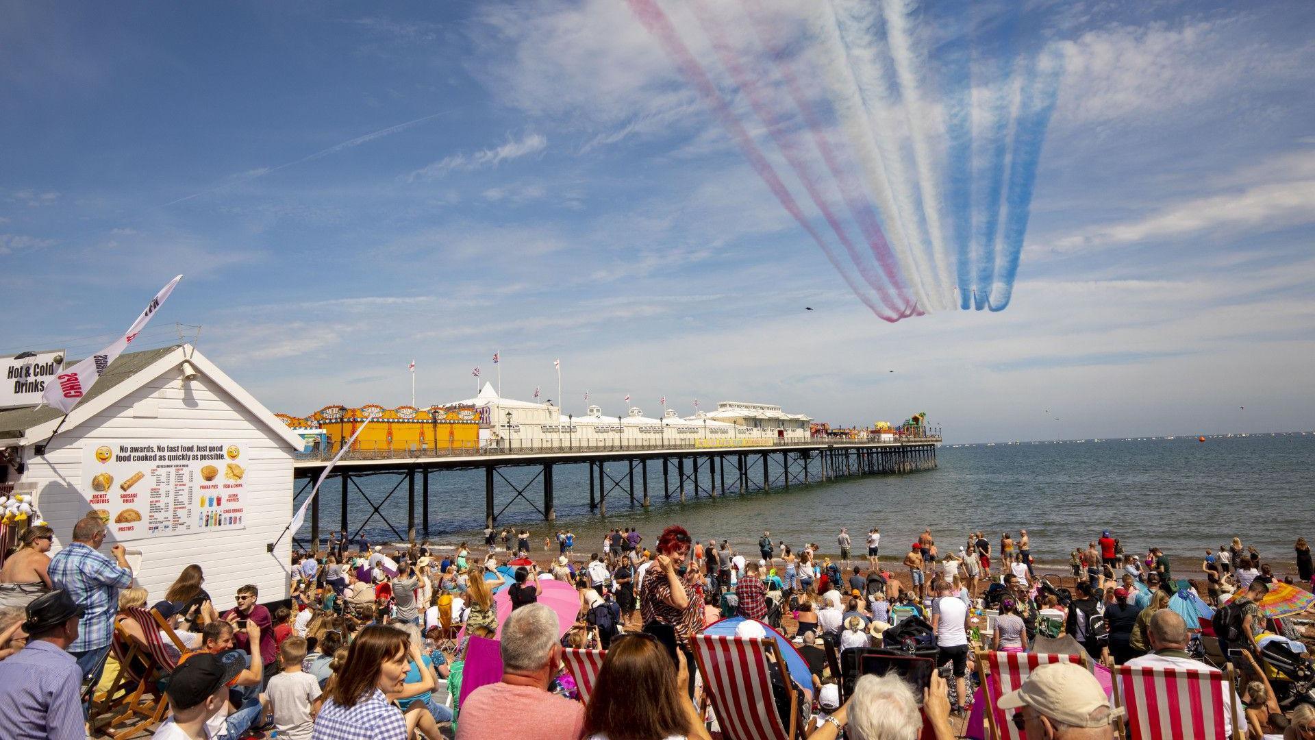 Crowds of people sat on Paignton beach admiring a Red Arrows display overhead, the blue sea in the distance alongside Paignton pier, on a sunny day.