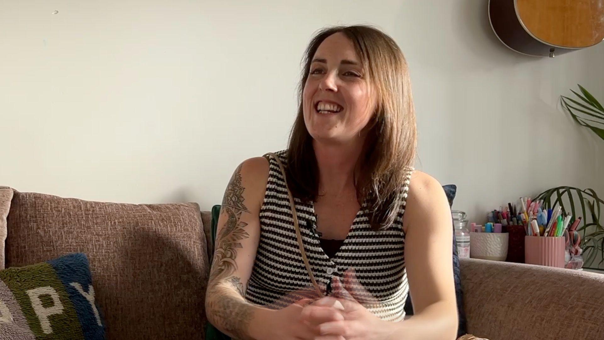 Sammy is sitting on a brown sofa. She is wearing a white and black striped sleevless top. She has medium length brown hair.