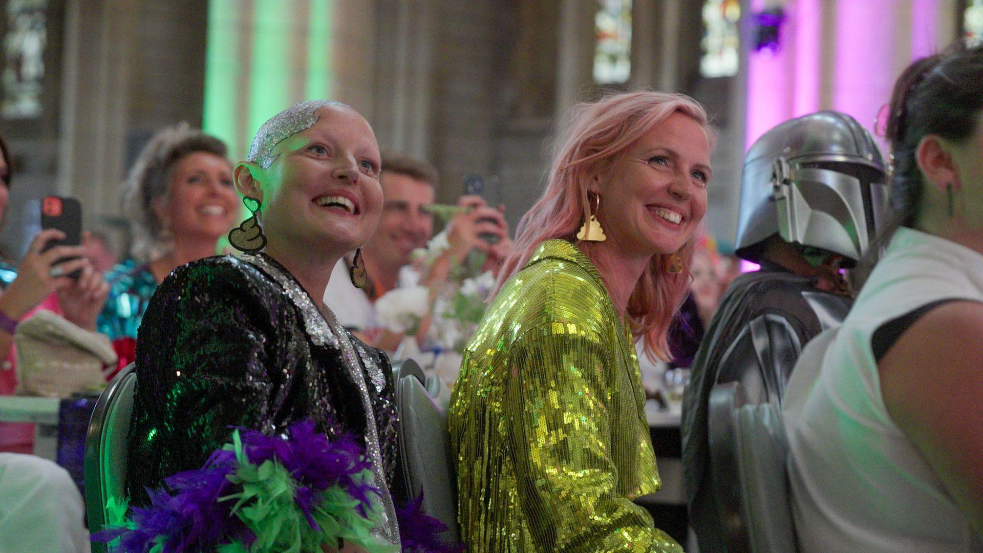 Kris and Maren are sitting in the congregation smiling. Kris is wearing a black and silver sparkly jacket with red and green feathers on it. Maren is wearing a green sparkly jacket. Sitting besides Maren is a person dressed in a suit of armour.