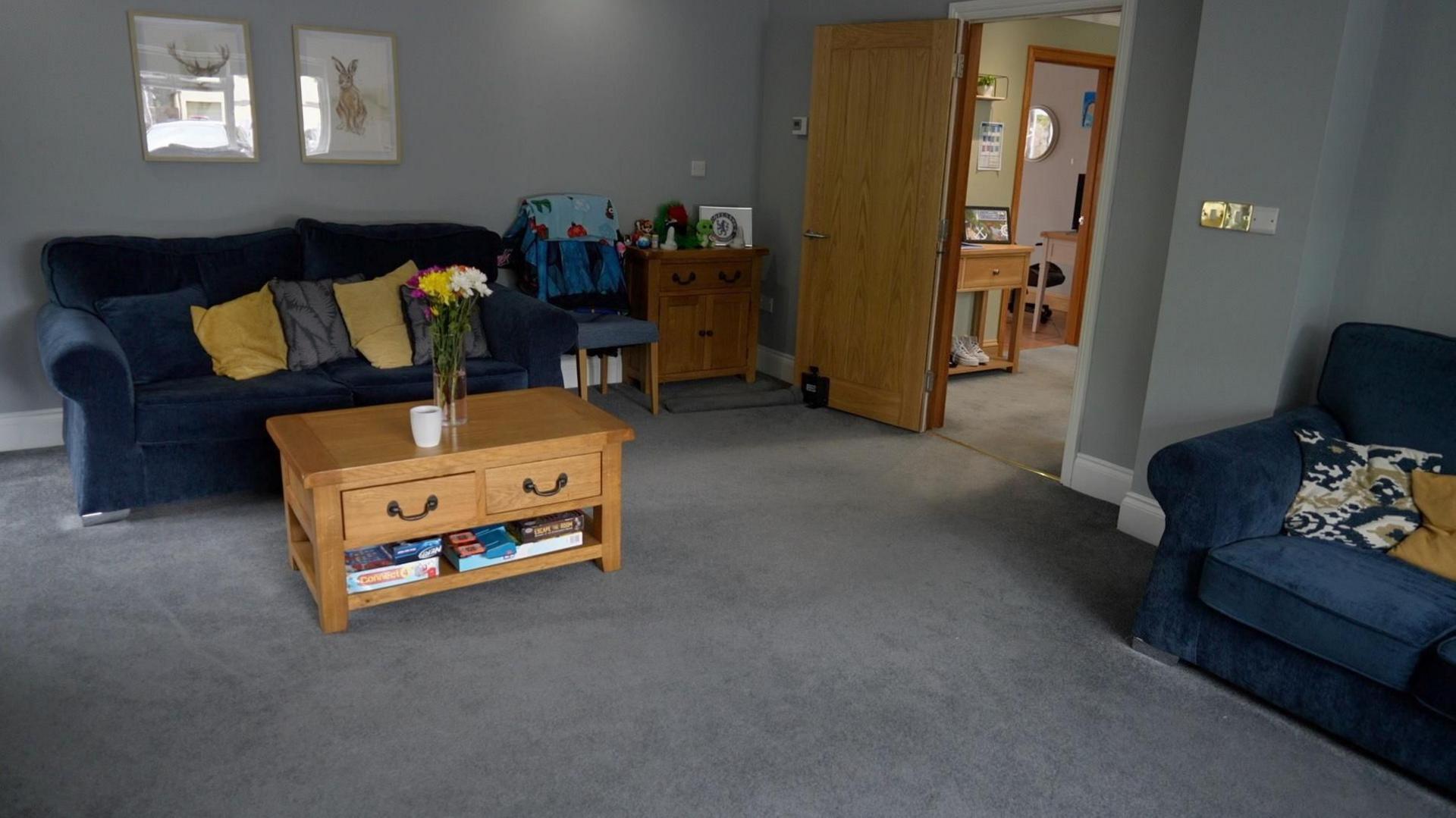 A living room with a grey carpet and two navy sofas with yellow accent cushions plus a pine coffee table and sideboard. The walls are painted grey and there are framed pictures of deer on the wall