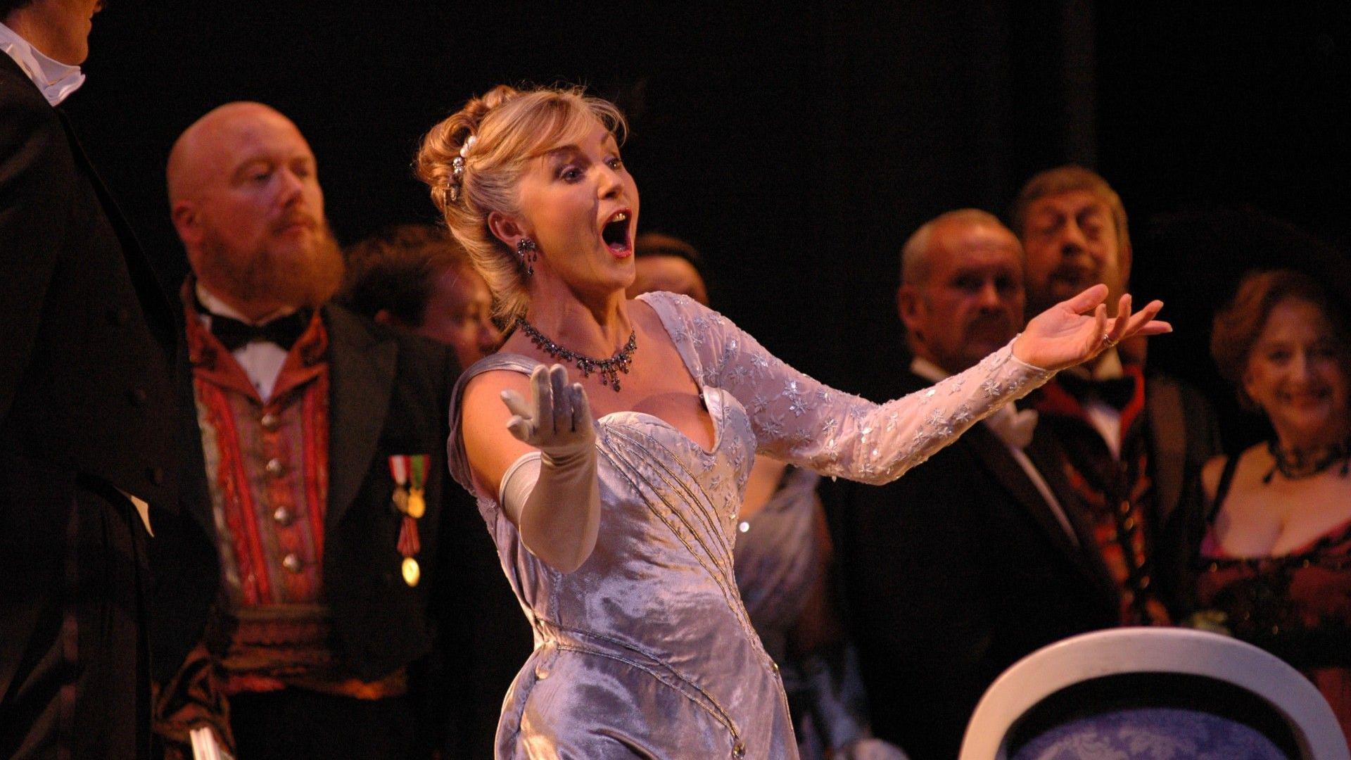 Lesley Garrett, singing in an opera, in Edwardian dress, with her hair up. Her mouth is open and her arms are out. Performers are behind her.