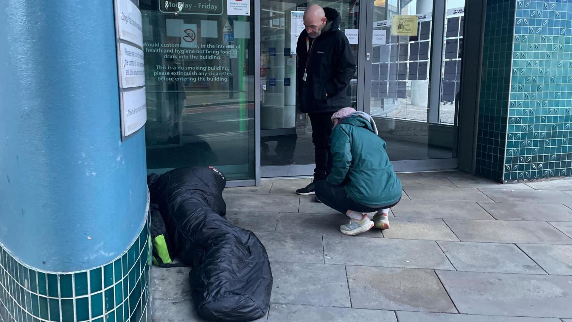 A woman in a green winter jacket and woolly hat squats down next to a man in a sleeping bag on the street. A second man stands looking down at them both.